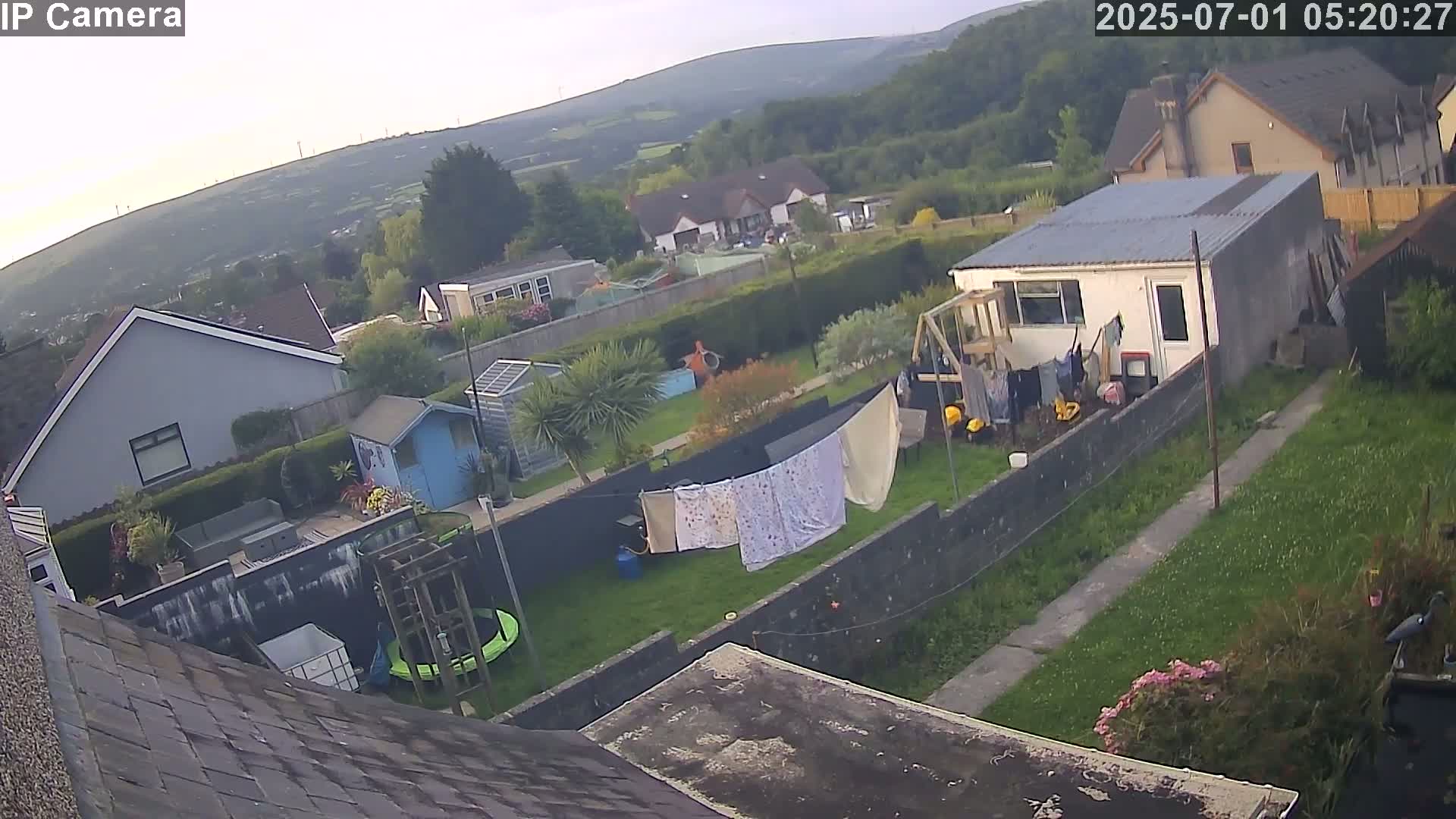 An overcast view of a residential backyard with a clothesline, small buildings, and a trampoline, overlooking a hillside of houses and greenery.