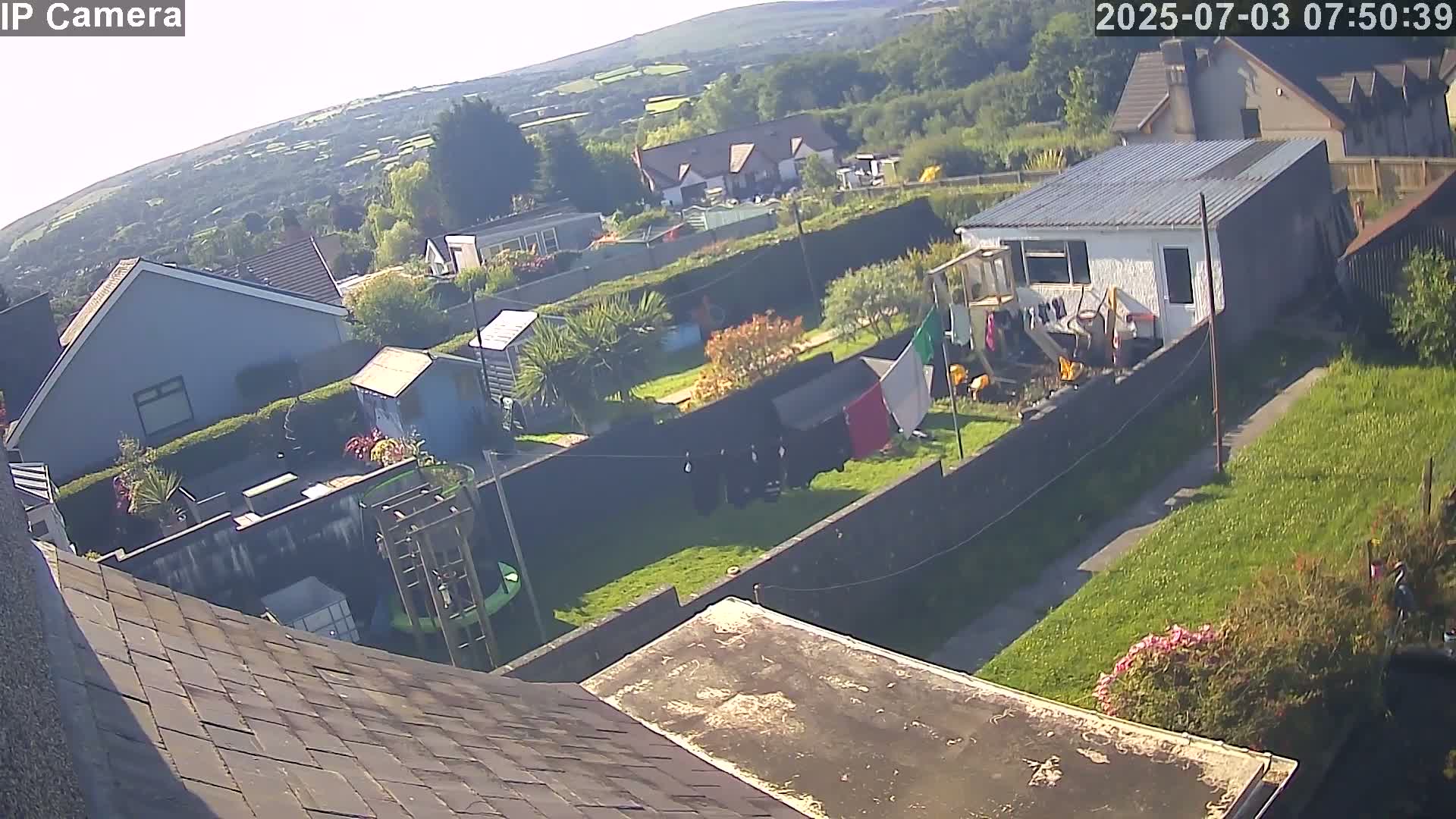 An aerial view shows a residential backyard with houses and greenery visible in the background under sunny conditions.