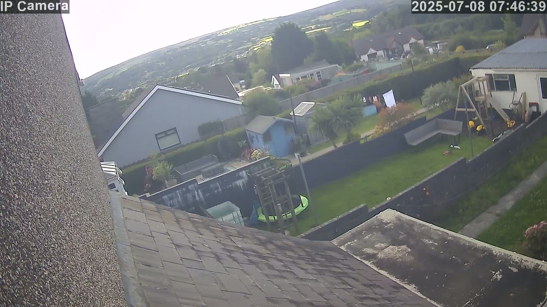 A high-angle view shows a residential backyard with a trampoline, playset, and small shed on a partly sunny day, overlooking a neighborhood and distant hills.