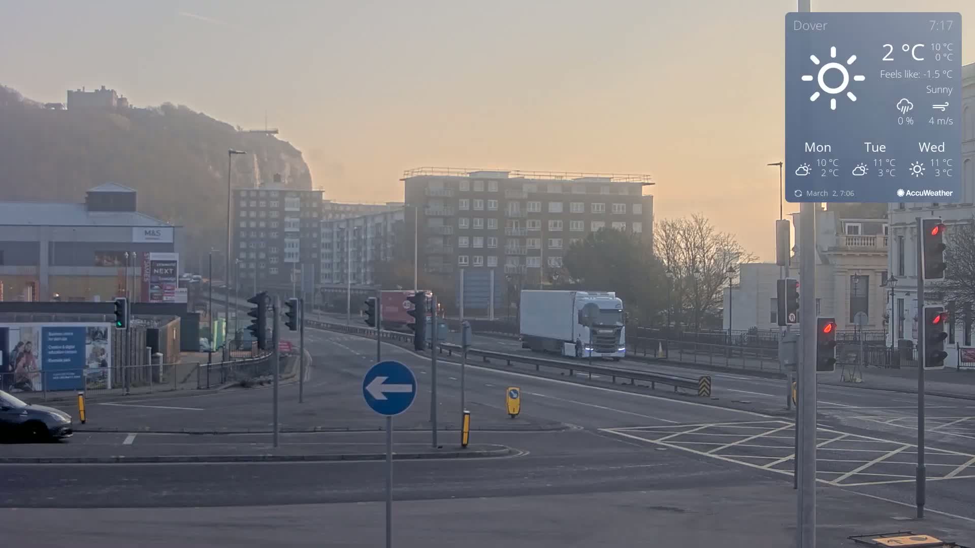 A sunny, hazy morning scene shows a multi-lane road with several vehicles, including a large white truck, passing through a traffic intersection near buildings and a hill with a castle in the distance.