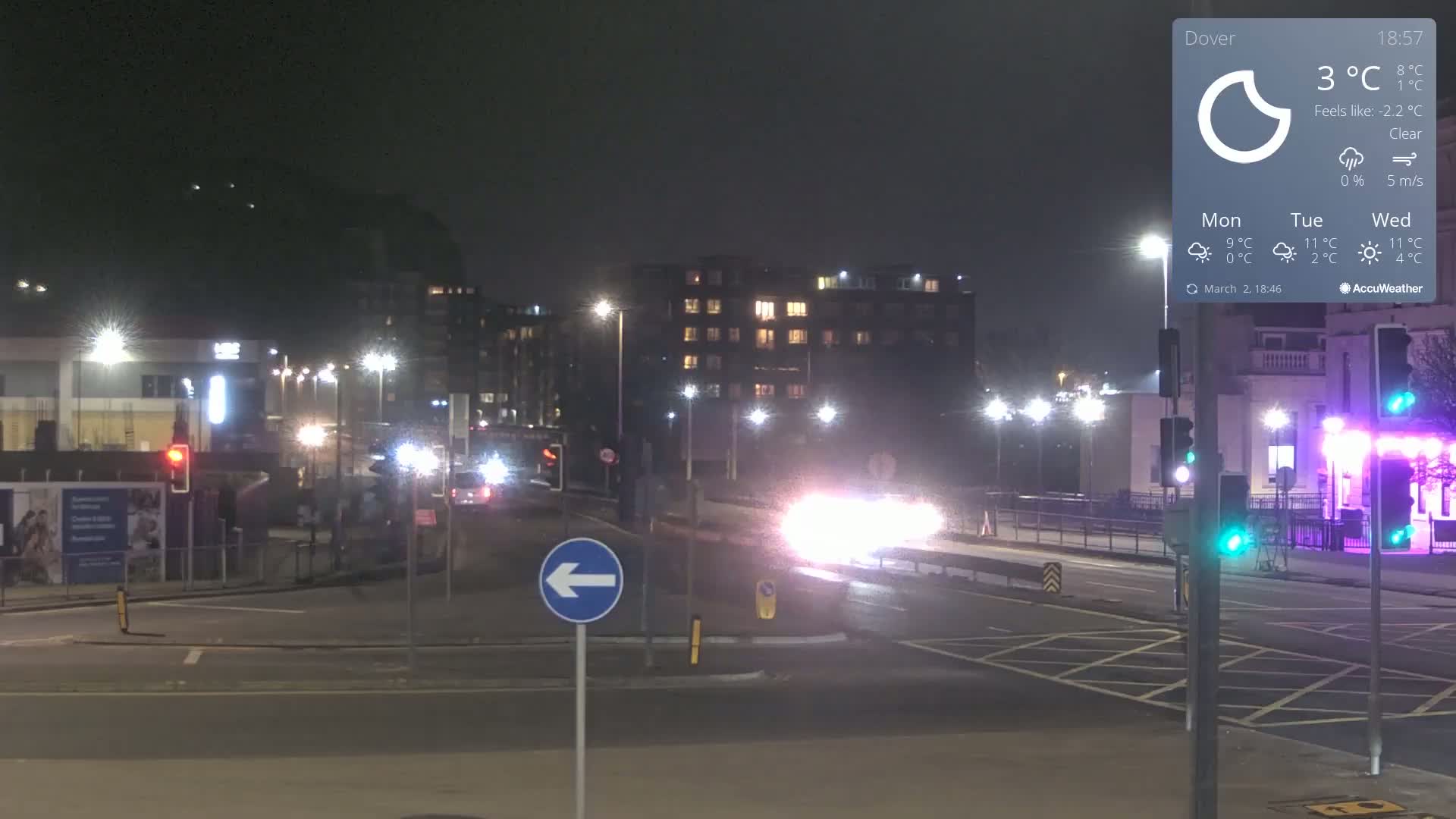 A nighttime street scene shows several vehicles traveling on a roadway, passing under streetlights in front of multi-story buildings, with clear skies and a temperature of 3°C.