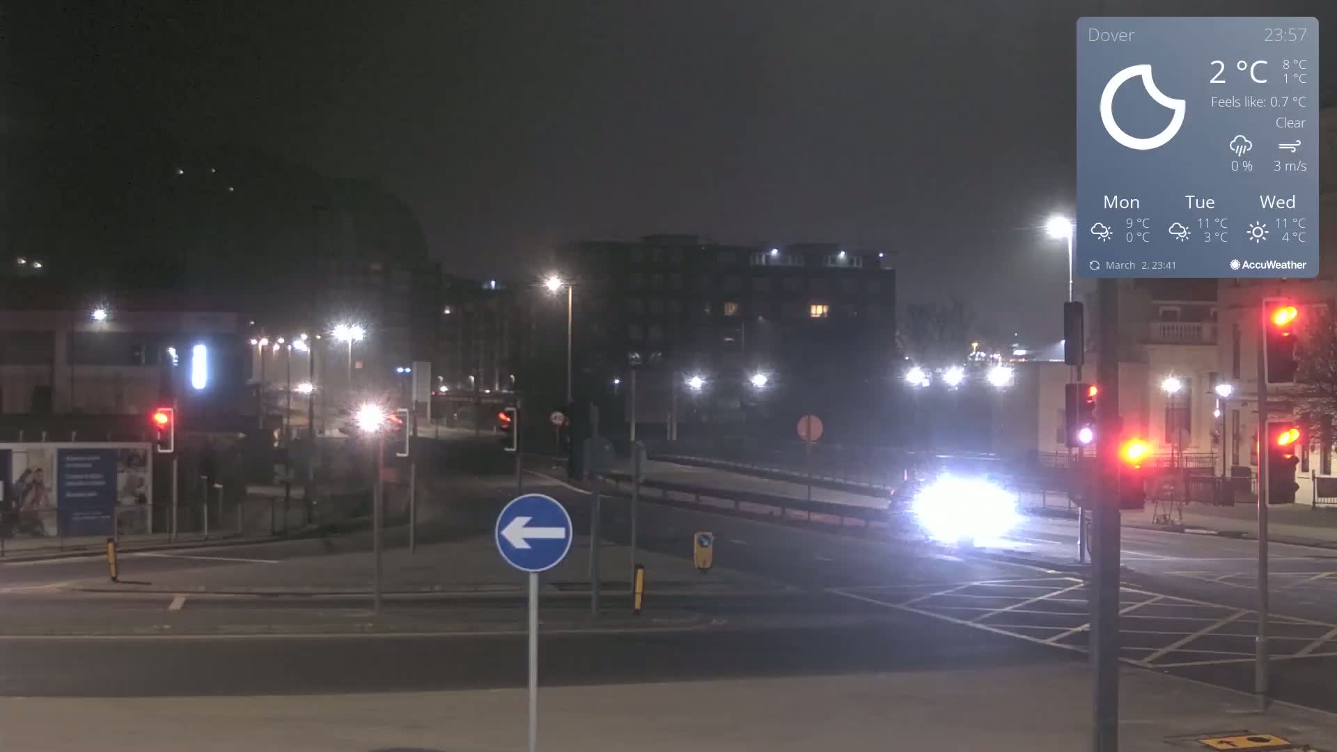A nighttime street scene shows a clear sky, a few cars on a roadway, and traffic lights at an intersection with buildings in the background.