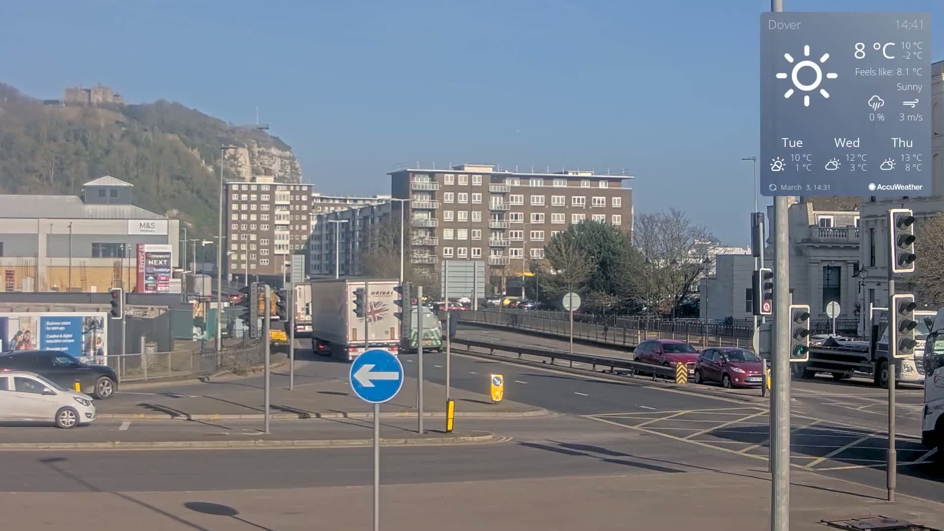 A sunny day shows a multi-lane intersection with several cars and trucks, buildings including a large apartment building, and a distant hilltop castle.