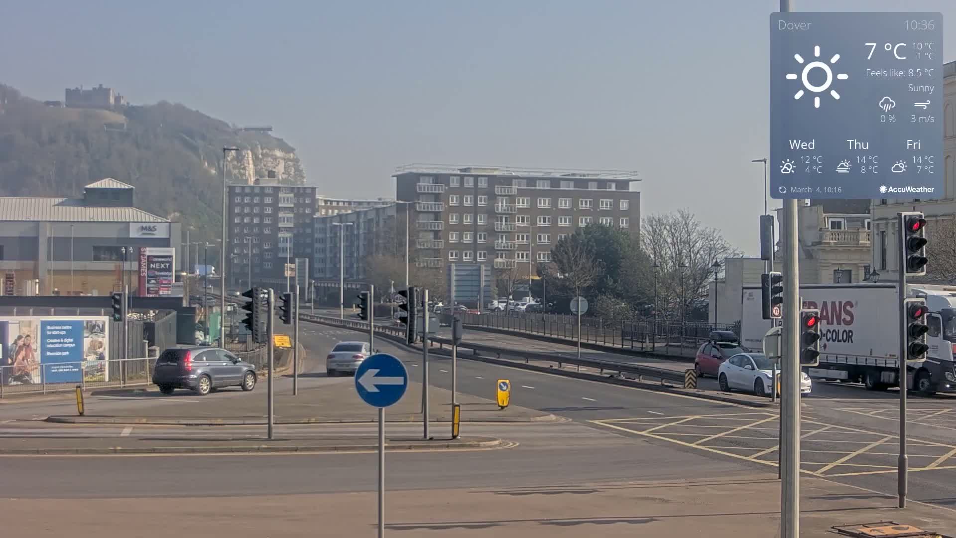 A sunny street scene shows several buildings, cars at an intersection, and a distant hilltop castle under a clear sky.