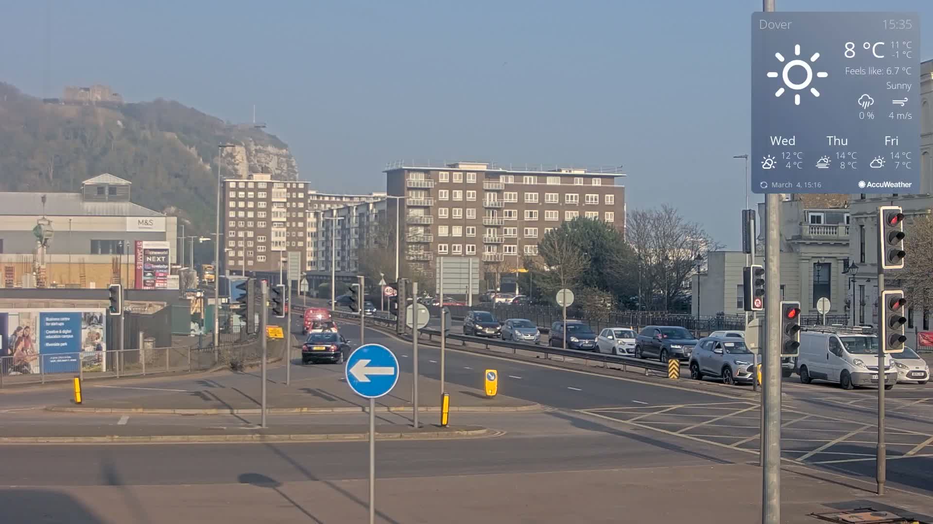 A sunny day shows a multi-lane road with cars and traffic signals in front of buildings and a hill in the background.