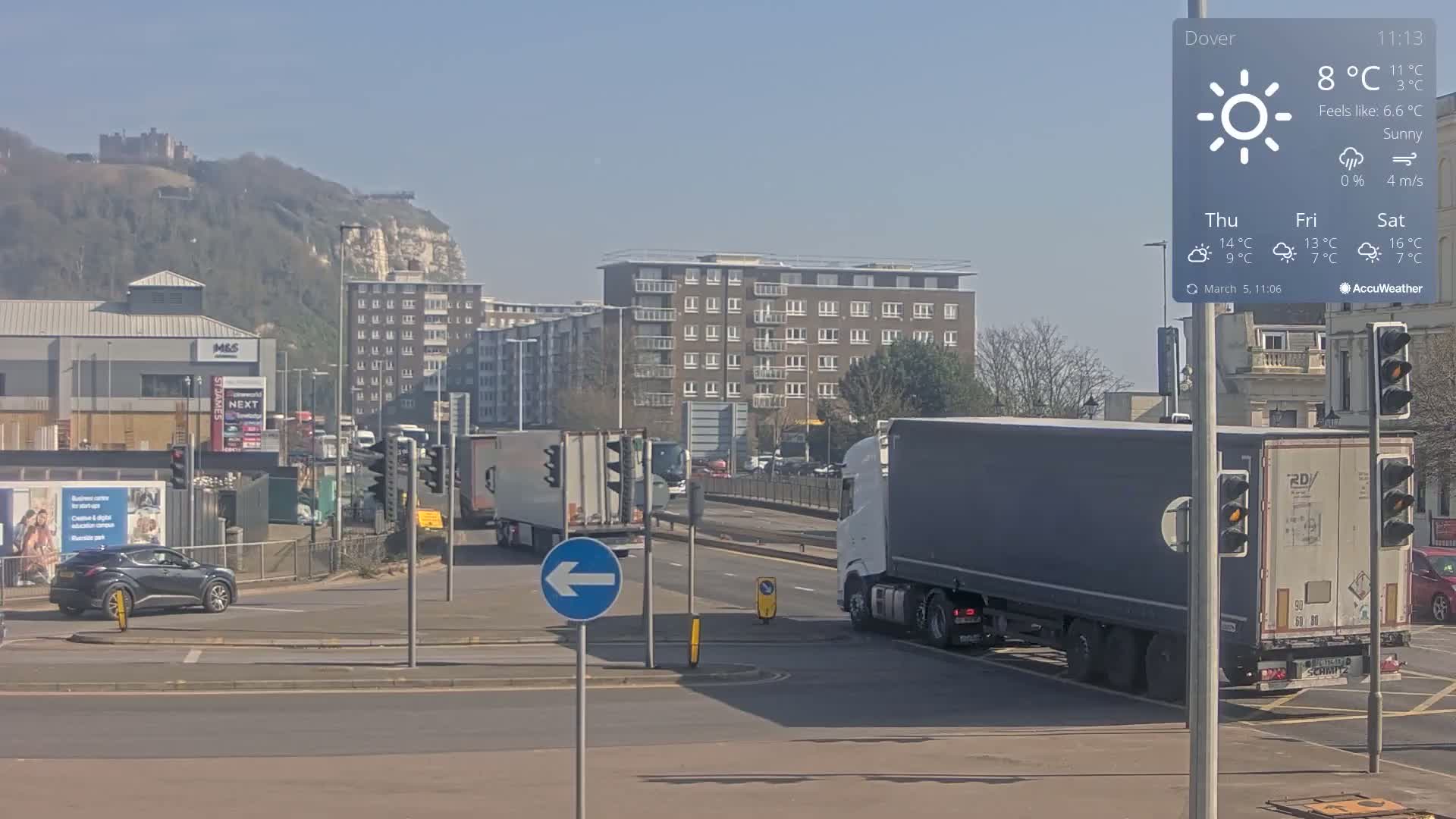 A sunny day shows a traffic intersection with several vehicles, including large trucks, and buildings in the background, with a castle visible on a hill in the distance.
