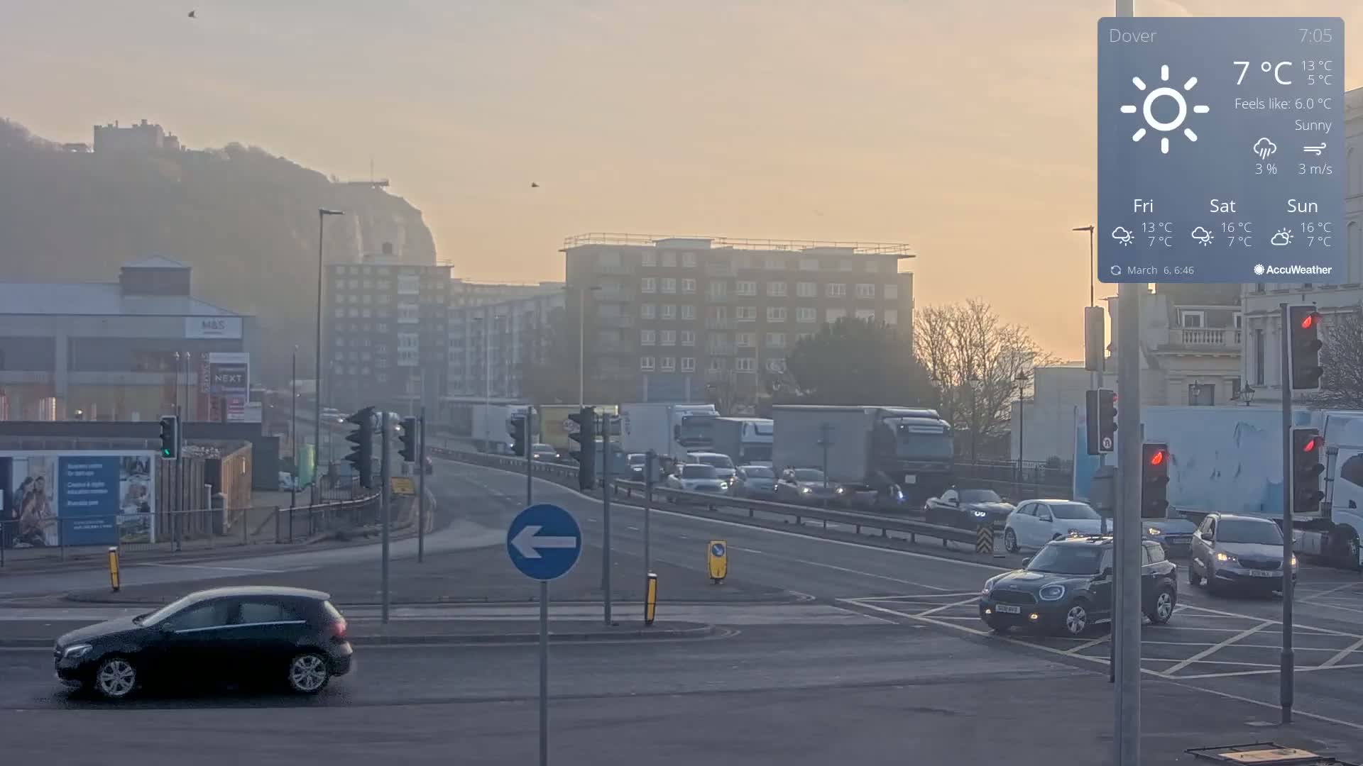 A hazy, sunny morning scene shows a busy intersection with cars and trucks, buildings, and a hill in the background.