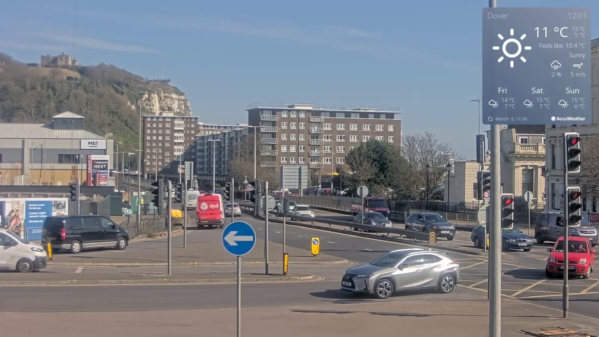 A sunny day reveals a busy intersection with multiple cars, a van, and buildings, including a multi-story apartment building, and a hill in the background showing a castle.