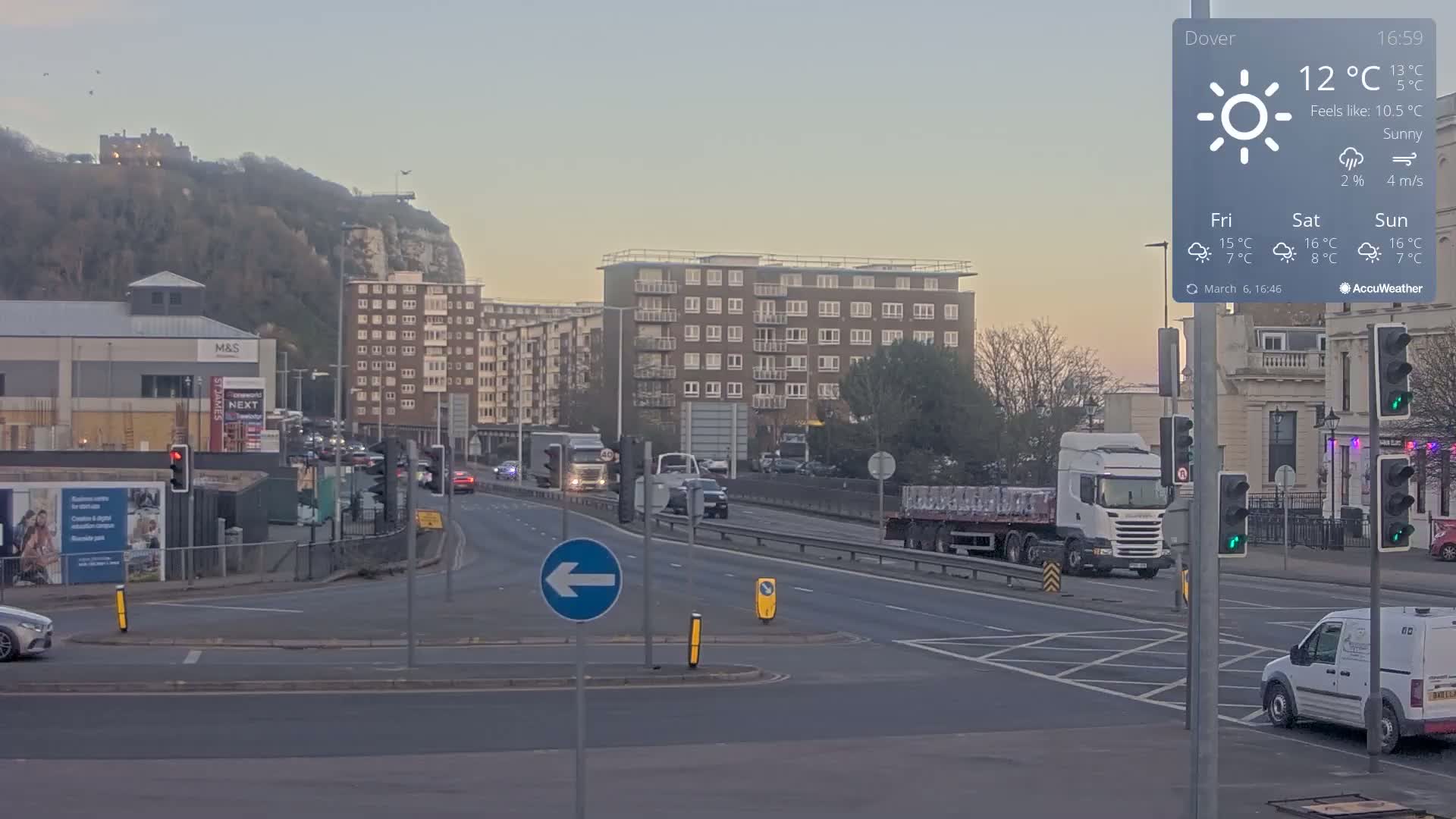 A sunny, yet cool day in a city with several buildings, a hill in the background, and traffic on a multi-lane road that has an intersection with traffic lights.