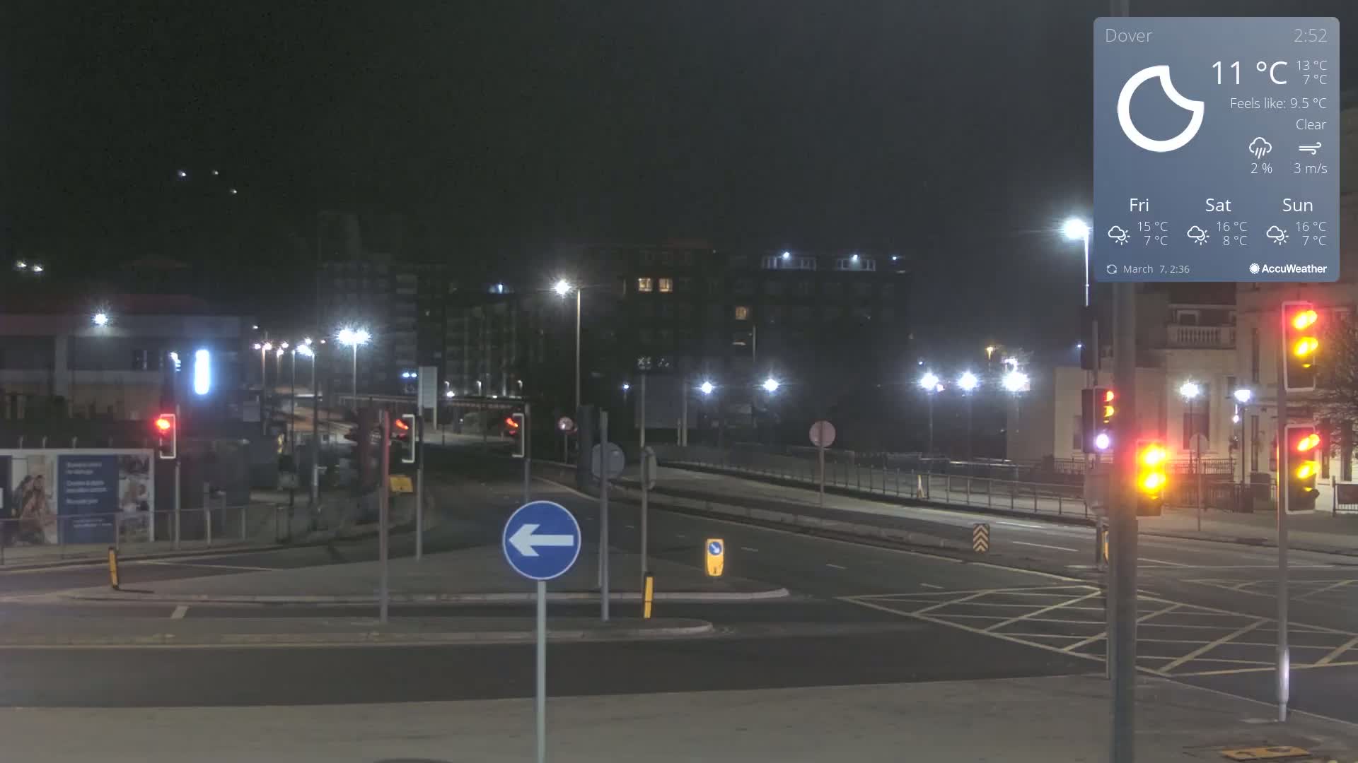 A nighttime view of a mostly empty intersection with traffic lights and streetlights illuminating buildings and a roadway under clear skies.