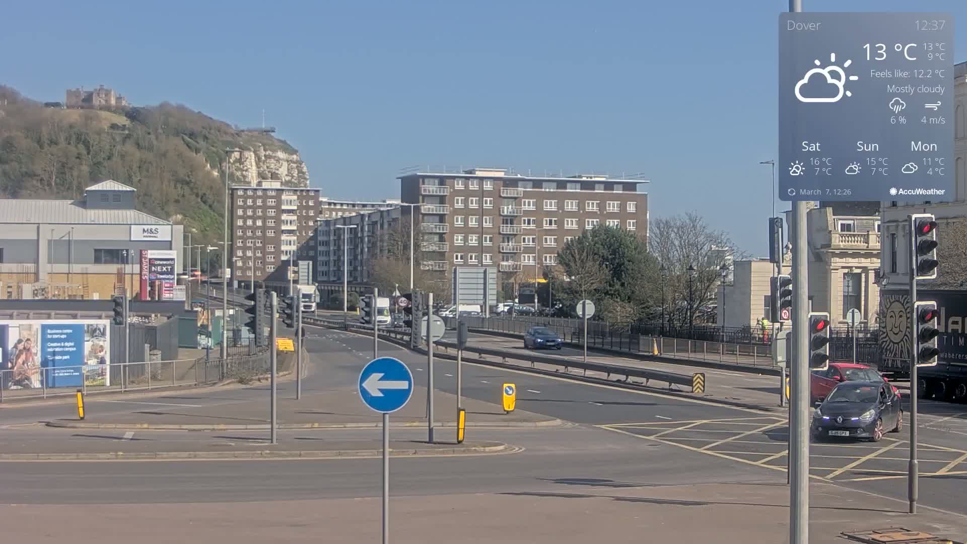 A mostly cloudy day reveals a city intersection with several cars, buildings, and a distant hill featuring a castle under a bright blue sky.