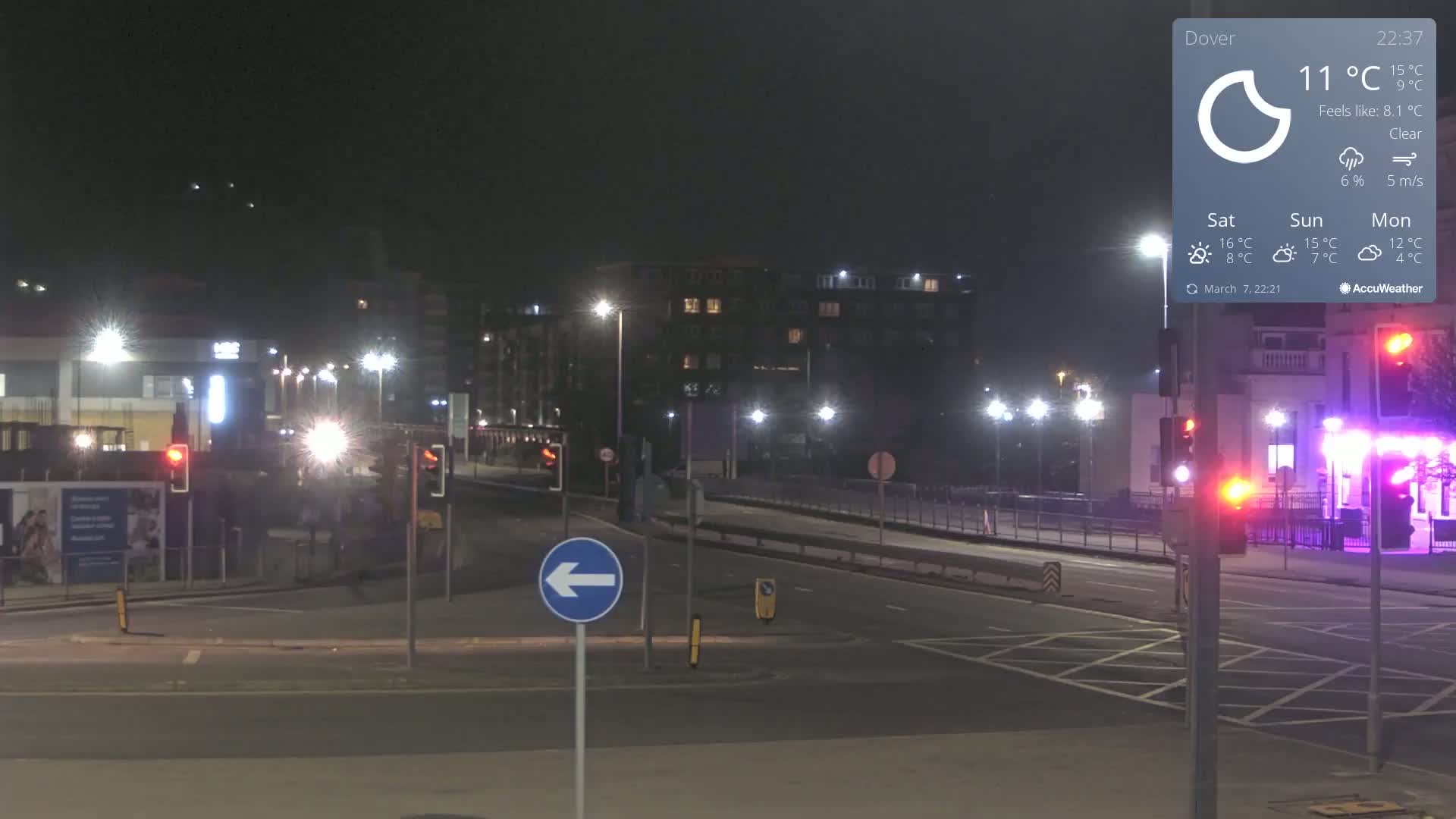 A nighttime street scene shows a mostly empty road with traffic lights and buildings in the background under clear skies.