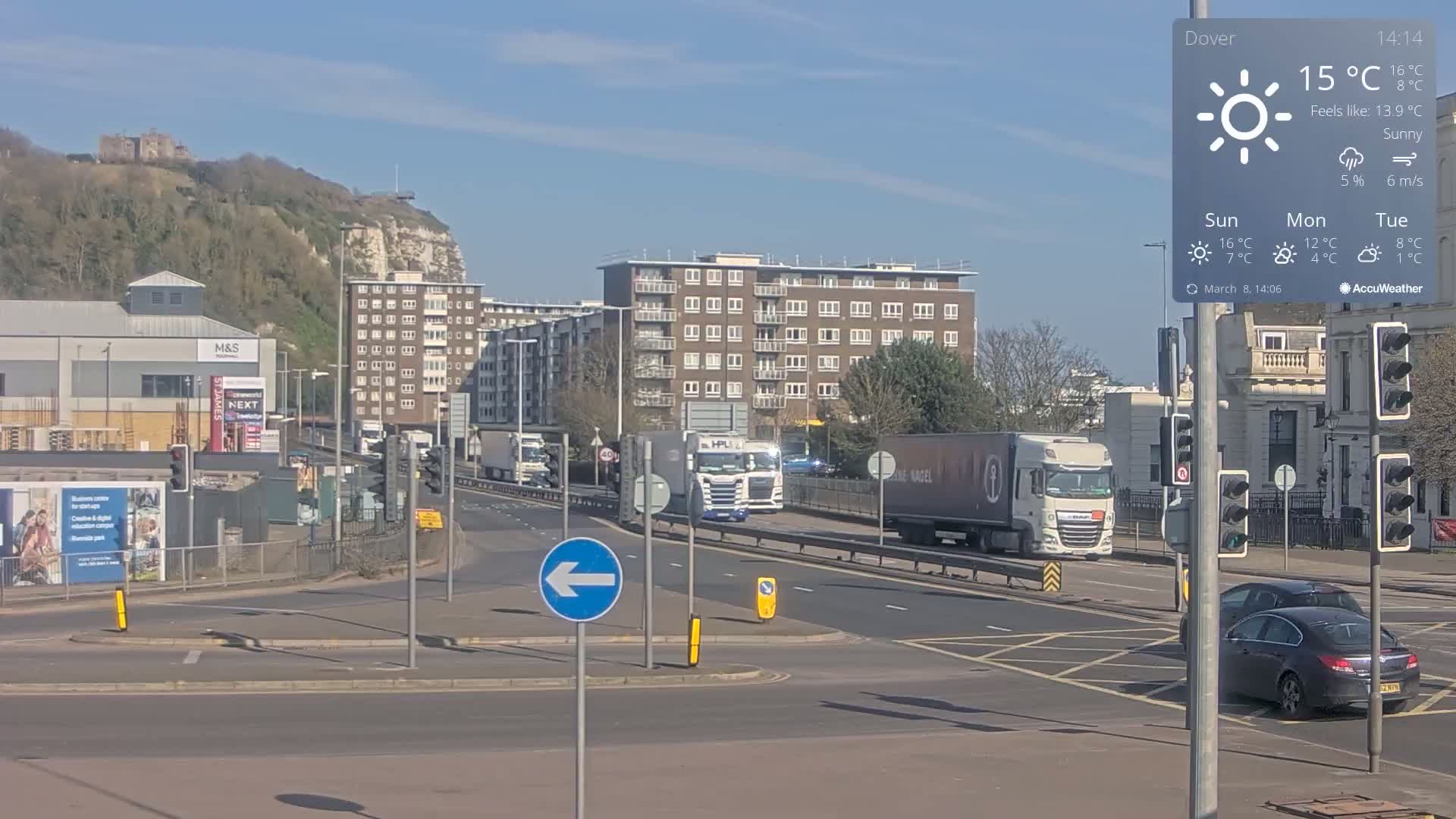 Under sunny skies, several large trucks and cars navigate a multi-lane roadway near a high-rise apartment building and a cliffside with a castle visible in the distance.