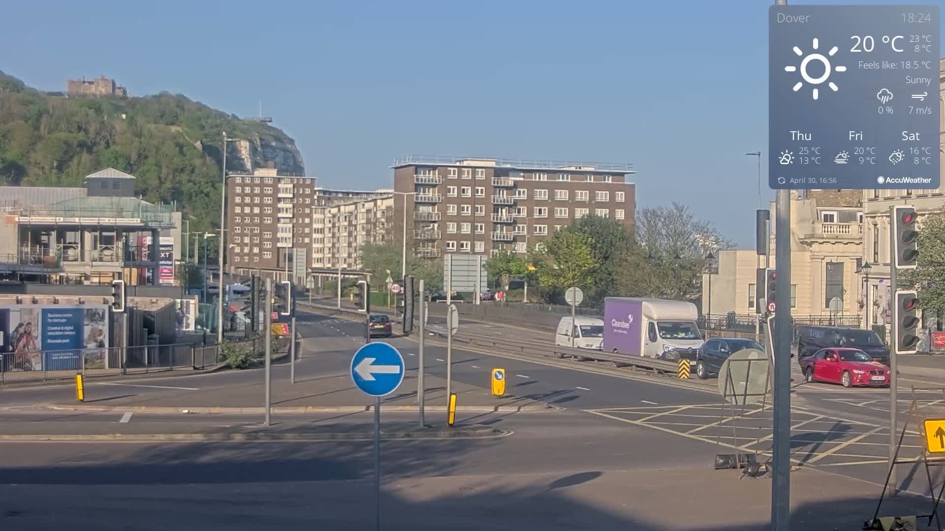 A sunny road intersection with several vehicles, buildings, and a distant castle on a hill.