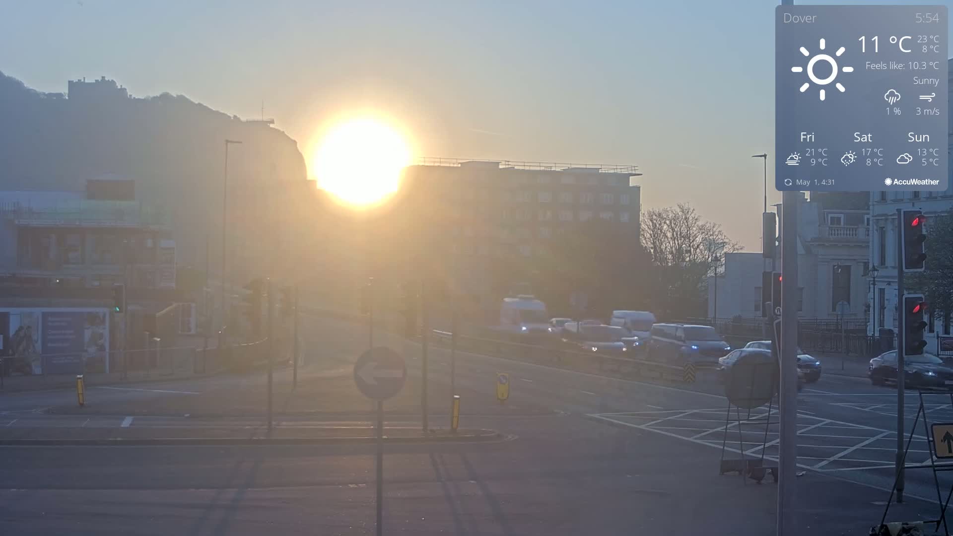 A hazy sunrise illuminates a street scene with cars at an intersection, a hill with a castle in the background, and buildings lining the road.
