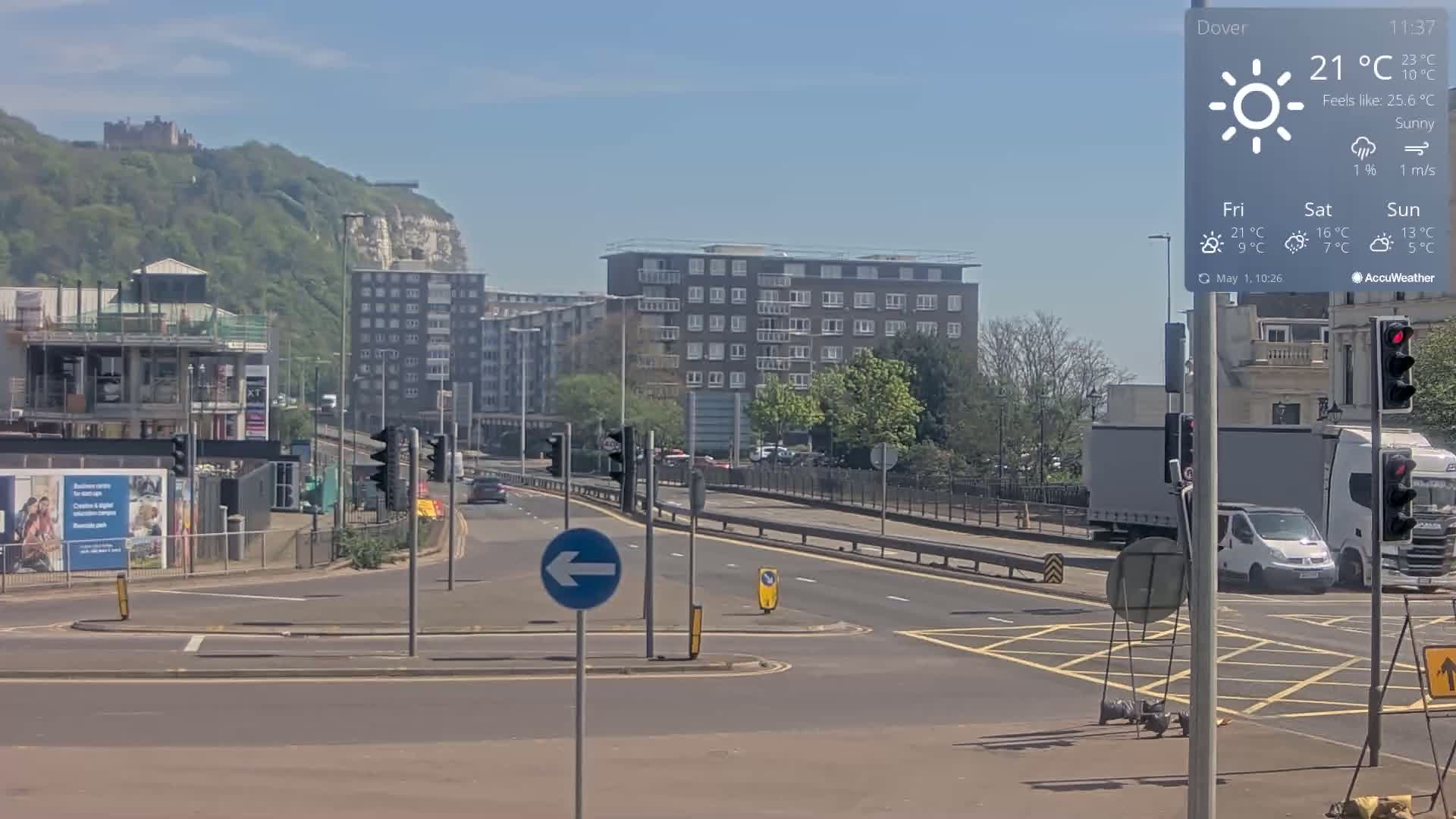 A sunny roadway with several cars and trucks intersects with a roundabout in front of multi-story apartment buildings and a hill topped with a castle.