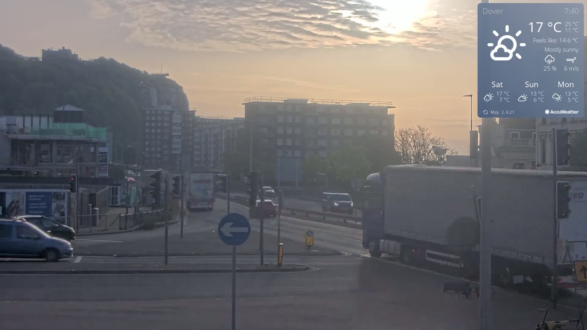 A mostly sunny morning reveals a seaside town with buildings, vehicles on a road, and a hill with a structure on top in the background.