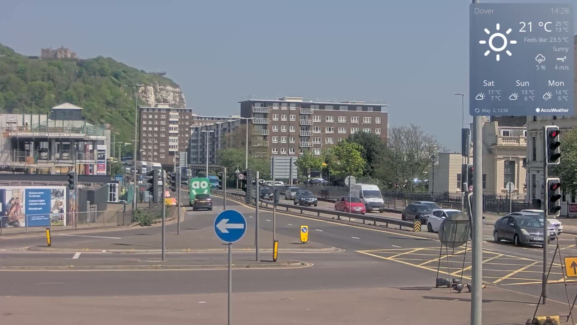 A sunny road intersection with multiple cars and a background of buildings, a hill, and a castle, under a clear sky.