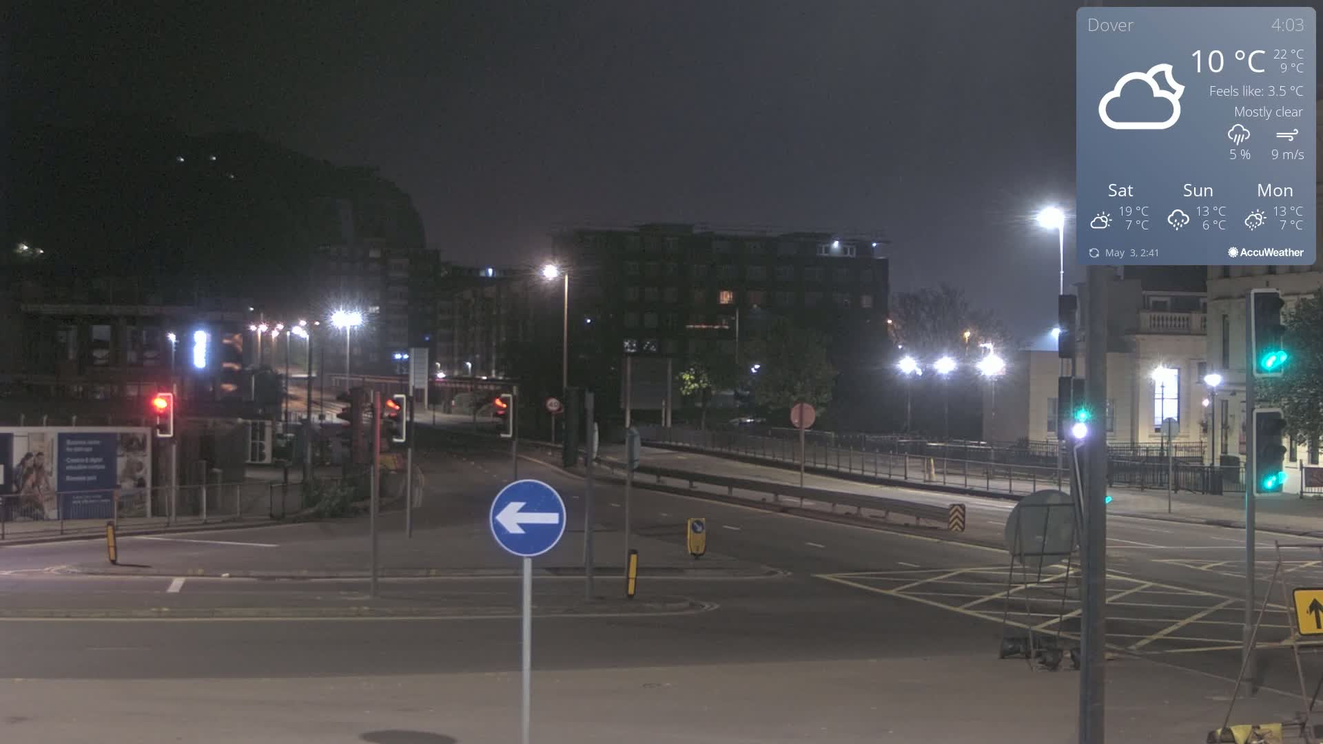 A mostly clear night scene shows a multi-lane road intersection with traffic lights and streetlights illuminating buildings and a hill in the distance.