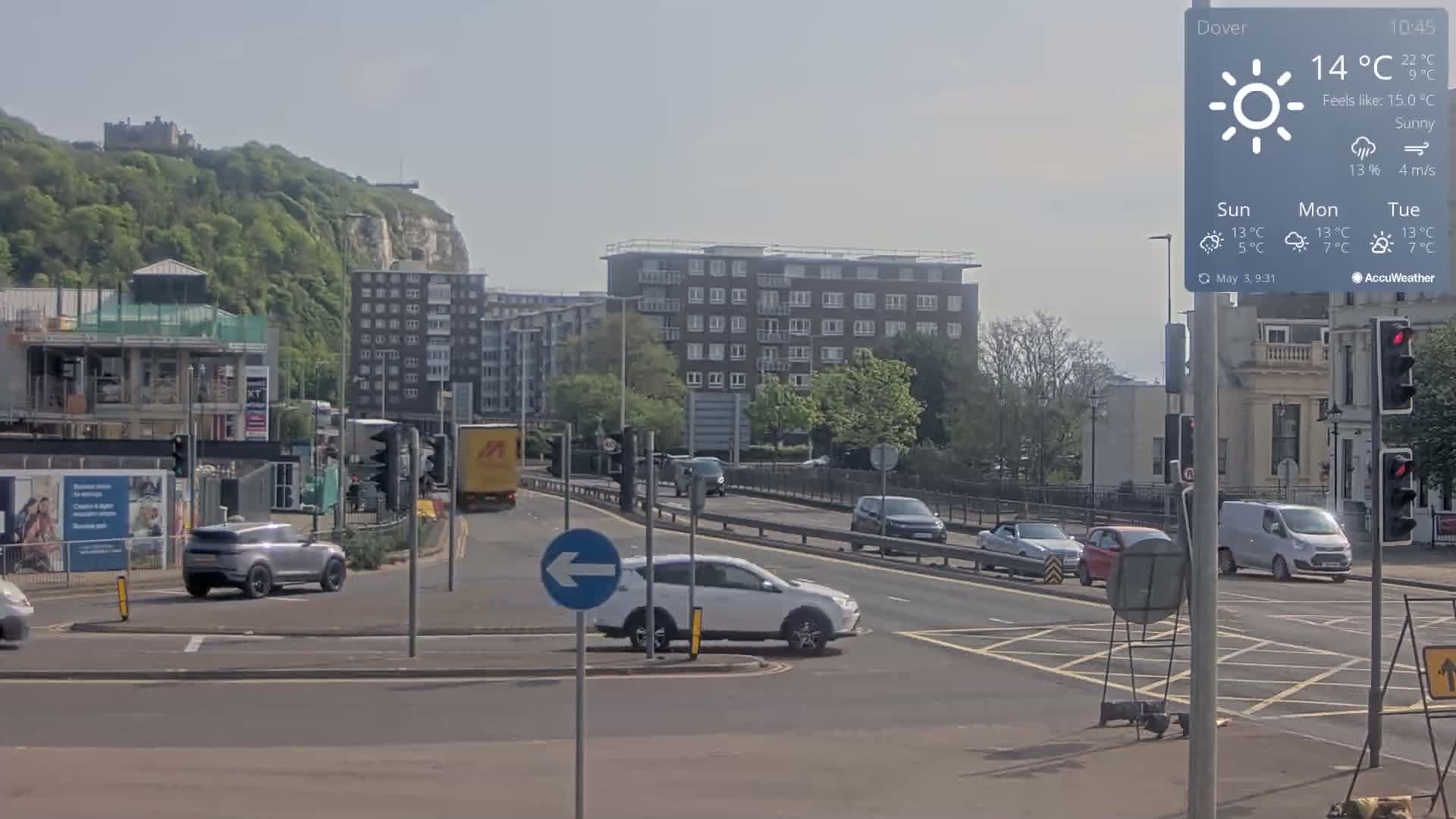 A sunny day shows several cars at a crossroads near a multi-story building and a hill with a castle visible in the distance.