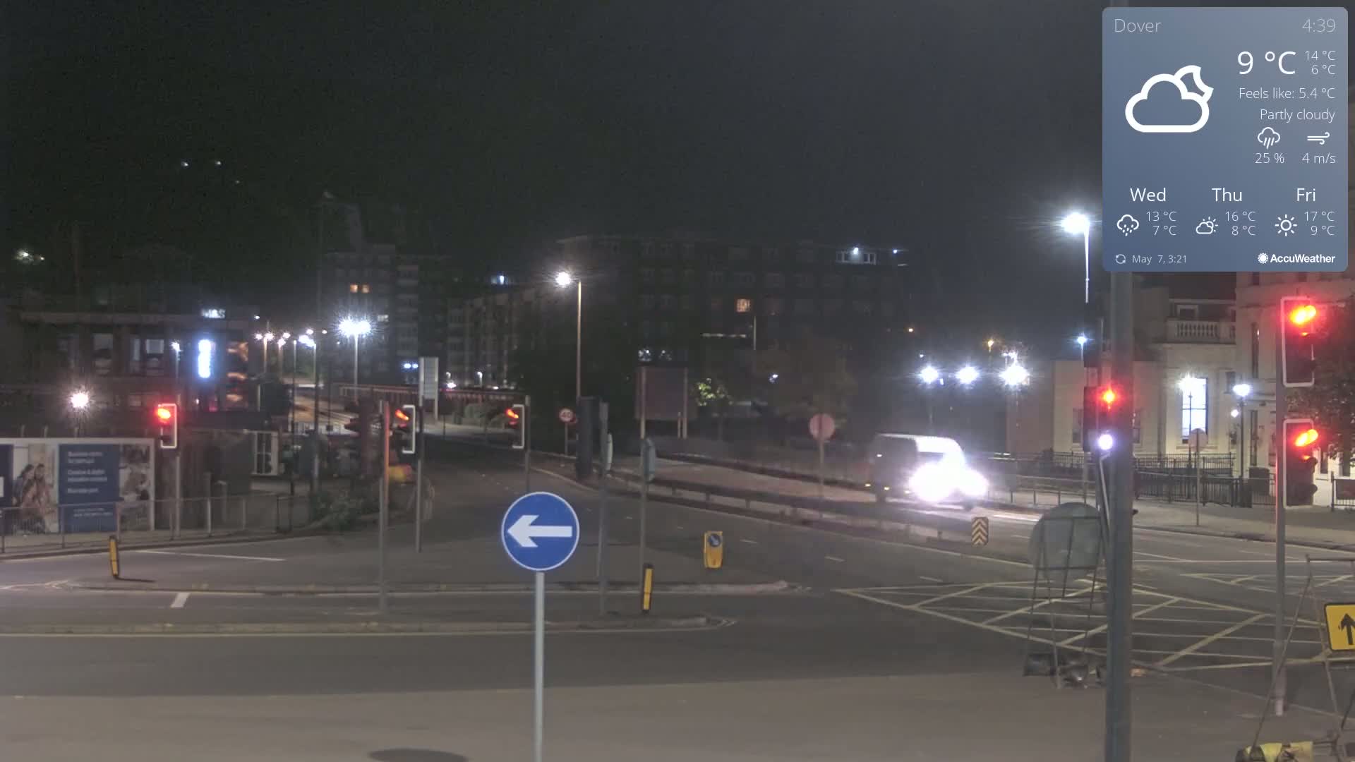 A nighttime, partly cloudy scene shows a mostly empty intersection with streetlights and a few vehicles under a partly cloudy sky.