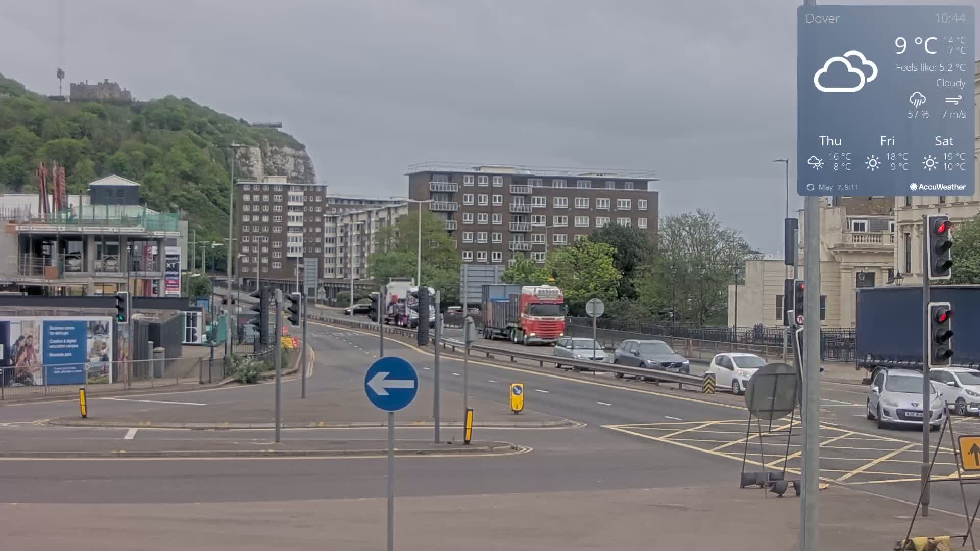 A cloudy day shows a road intersection with several cars and trucks, a large building under construction, and apartment buildings, all situated with a hill and castle in the background.