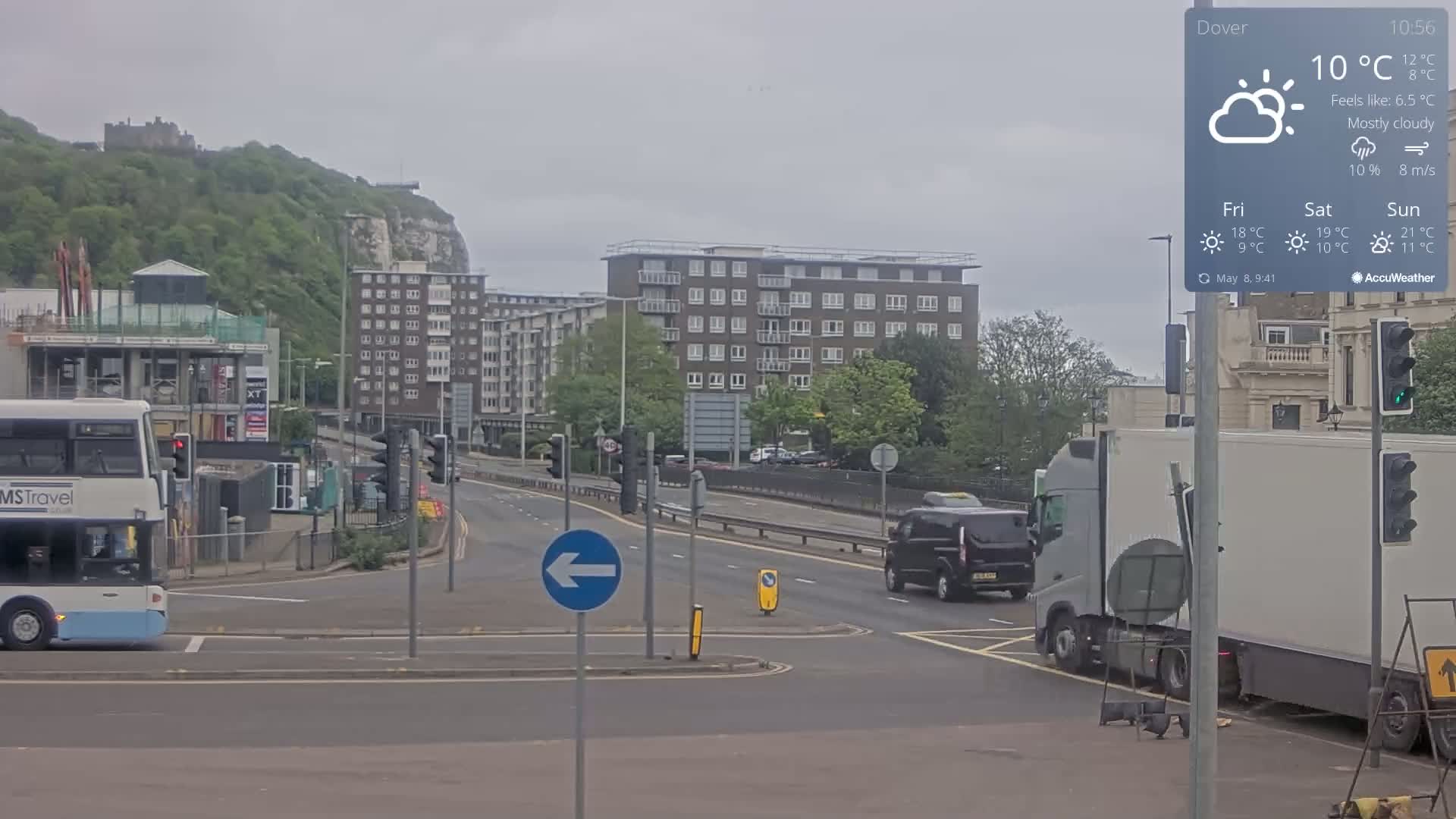A mostly cloudy day shows a street scene with a double-decker bus, a minivan, and a large truck at an intersection, near a hill with buildings and a castle in the distance.