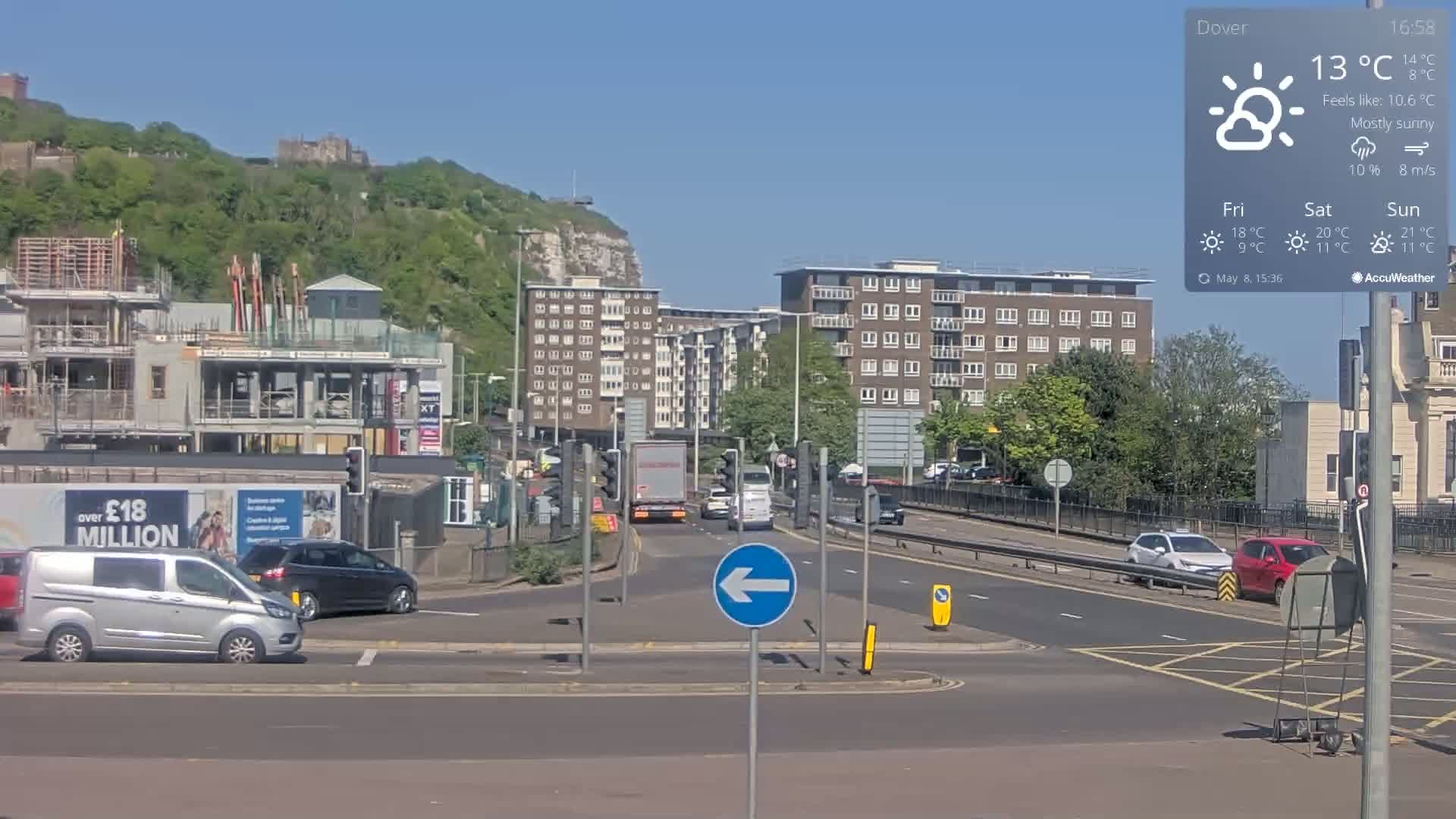 A mostly sunny scene shows a busy intersection with several cars and trucks, construction sites, apartment buildings, and a hill with a castle in the background.