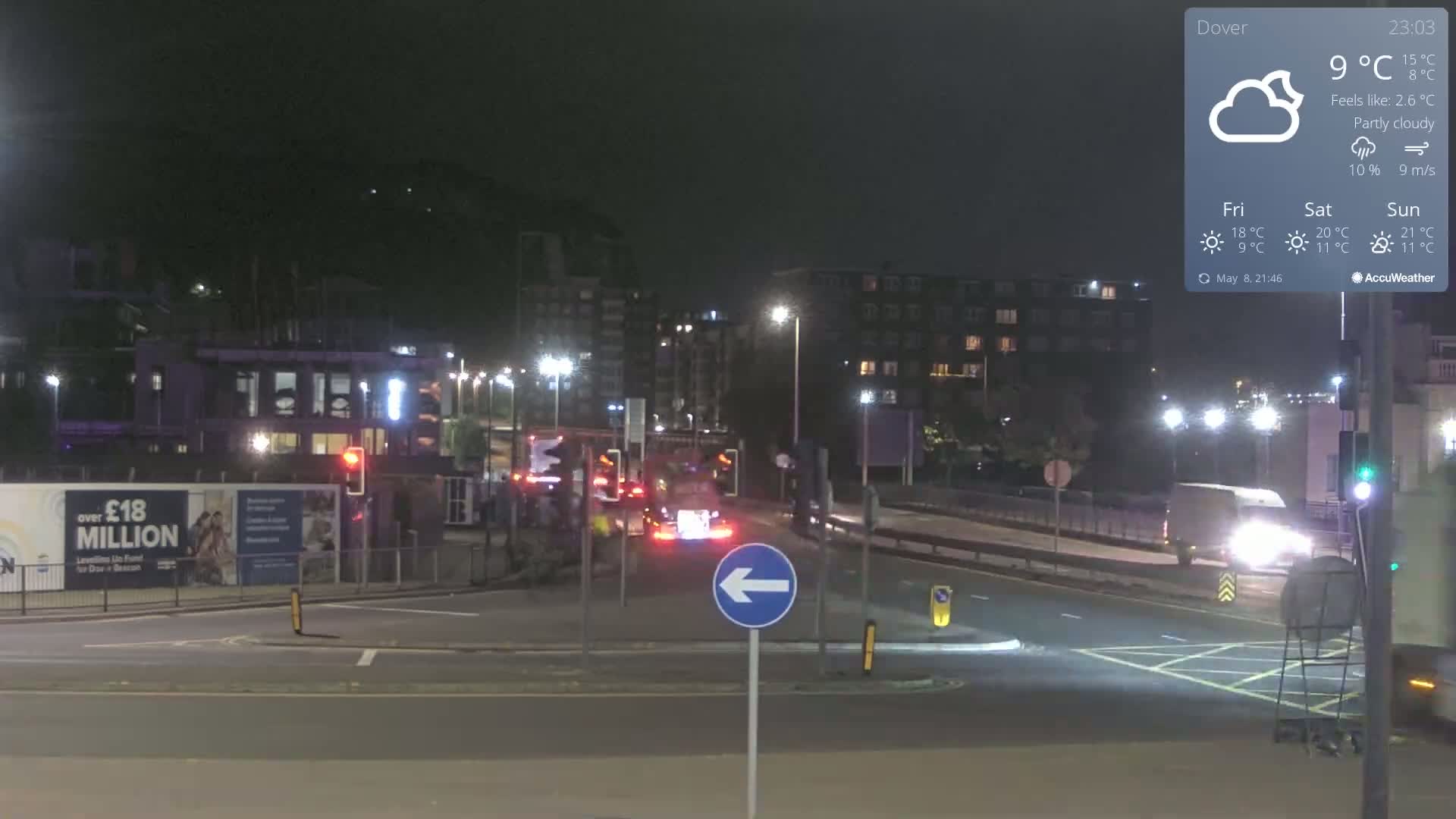 A nighttime, partly cloudy scene shows a traffic intersection with several vehicles, buildings in the background, and a blue left-turn arrow sign.