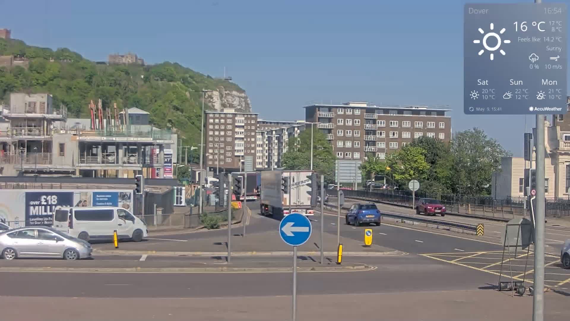 A sunny, 16°C day reveals a city street scene with several vehicles at an intersection, construction sites, apartment buildings, and a hill topped with a castle in the background.