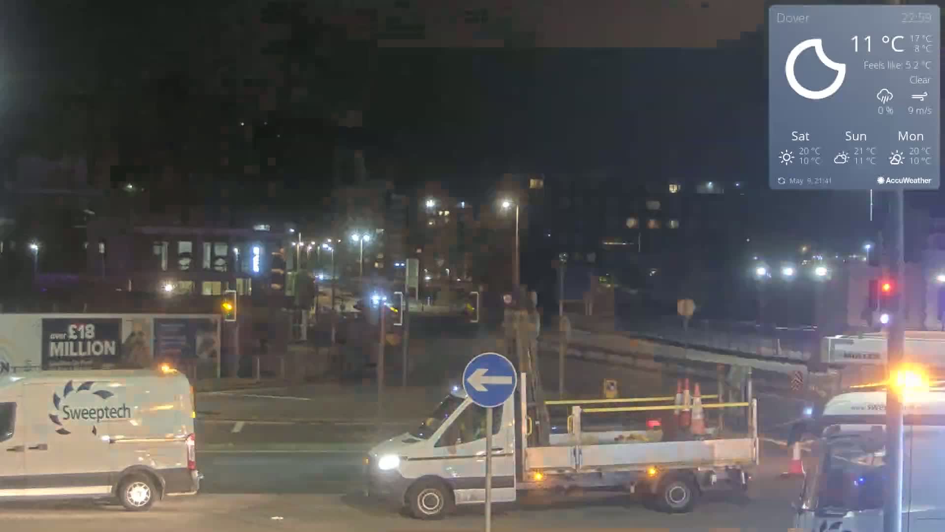 At night, under clear skies, a white van and a flatbed truck are stopped at an intersection, with other vehicles and buildings visible in the background.