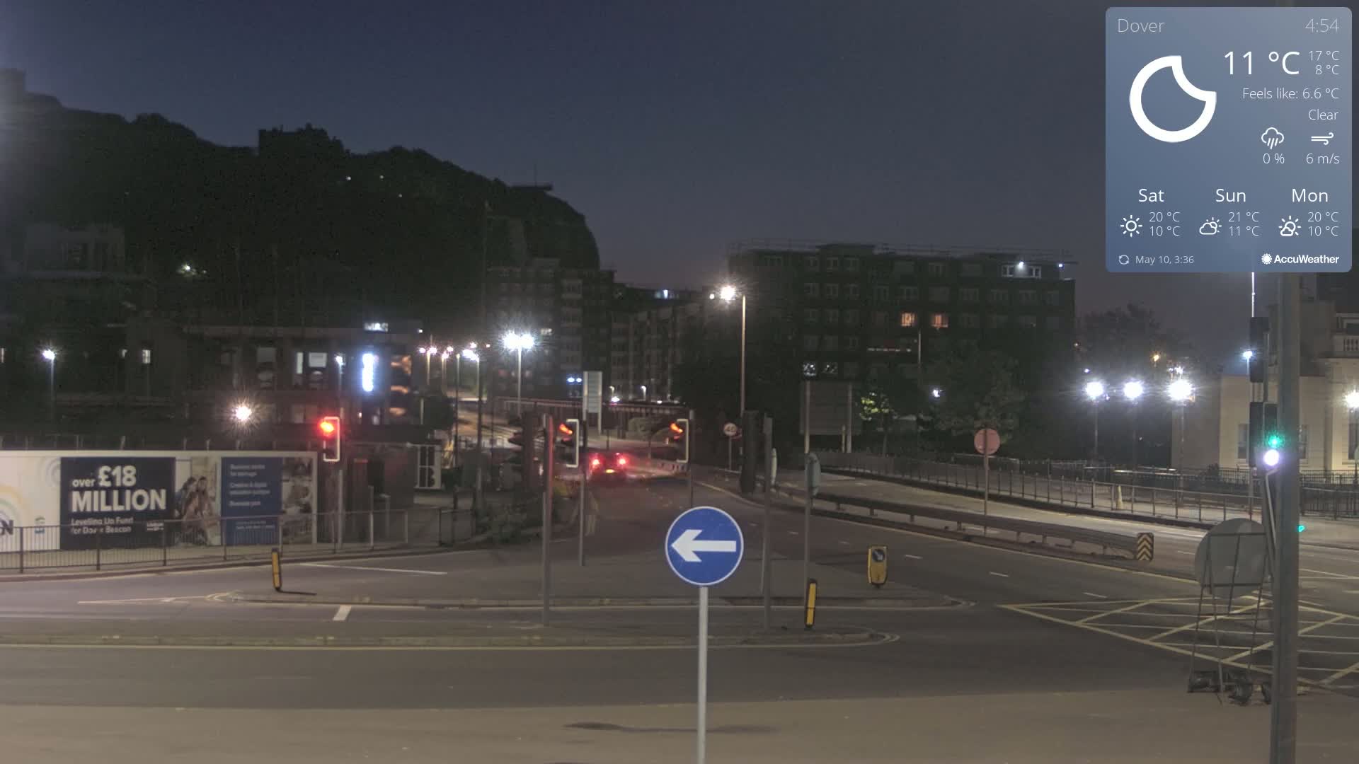 Under a clear night sky, a mostly empty roadway with streetlights and a few vehicles is shown, with buildings and a hill in the background.