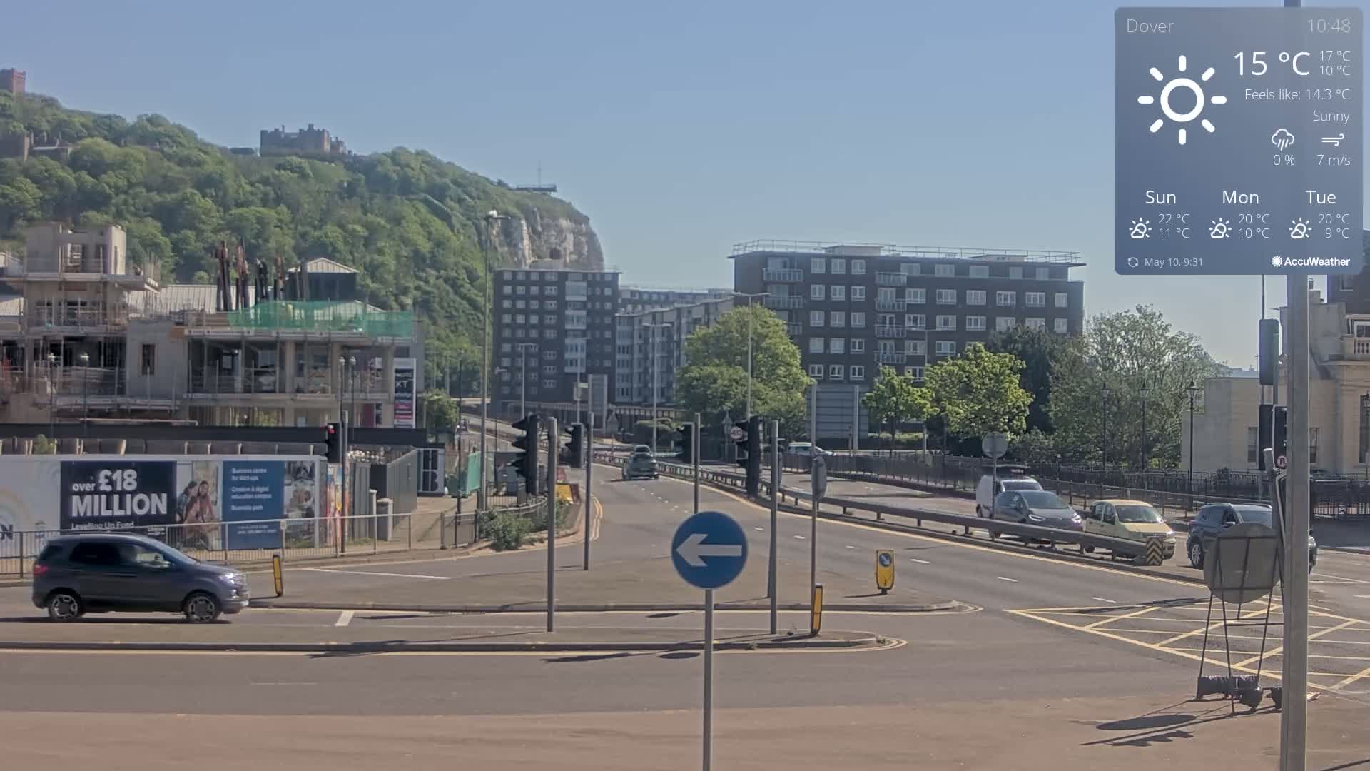 A sunny city street scene shows several cars driving on a road that passes under a traffic light and near a construction site with a partially built building, apartment buildings, and a castle visible on a hillside in the distance.