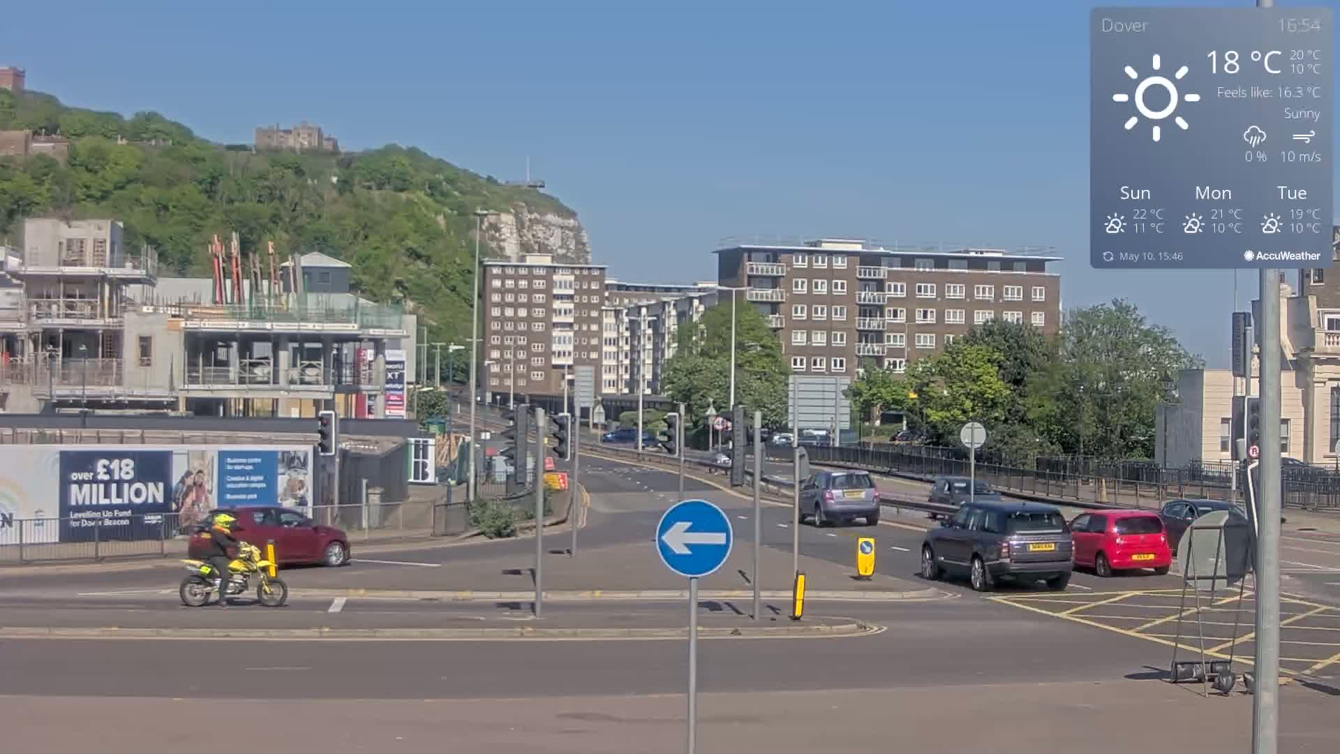 A sunny day shows a multi-lane road intersection with several cars and a motorcycle, near a construction site, apartment buildings, and a hill with greenery and a castle in the background.