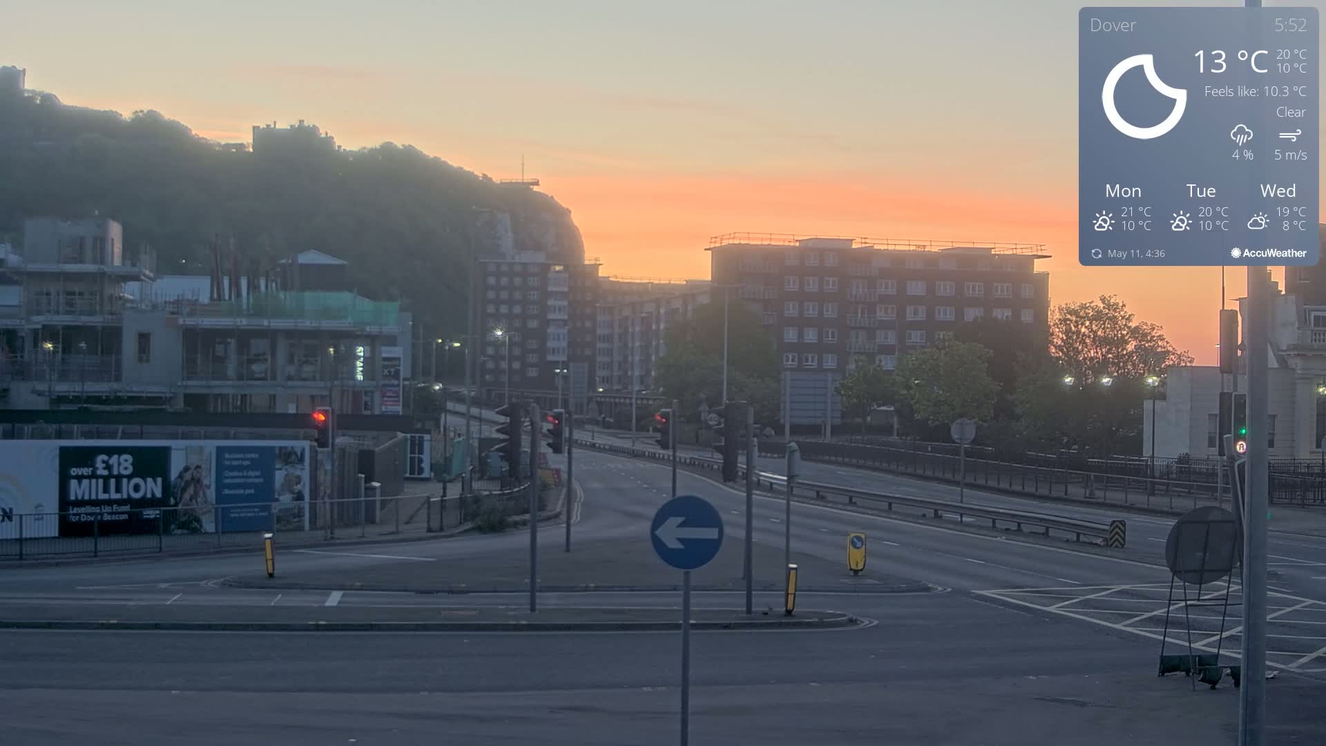 A mostly empty multi-lane road at sunrise, with buildings and construction sites visible under a clear sky.