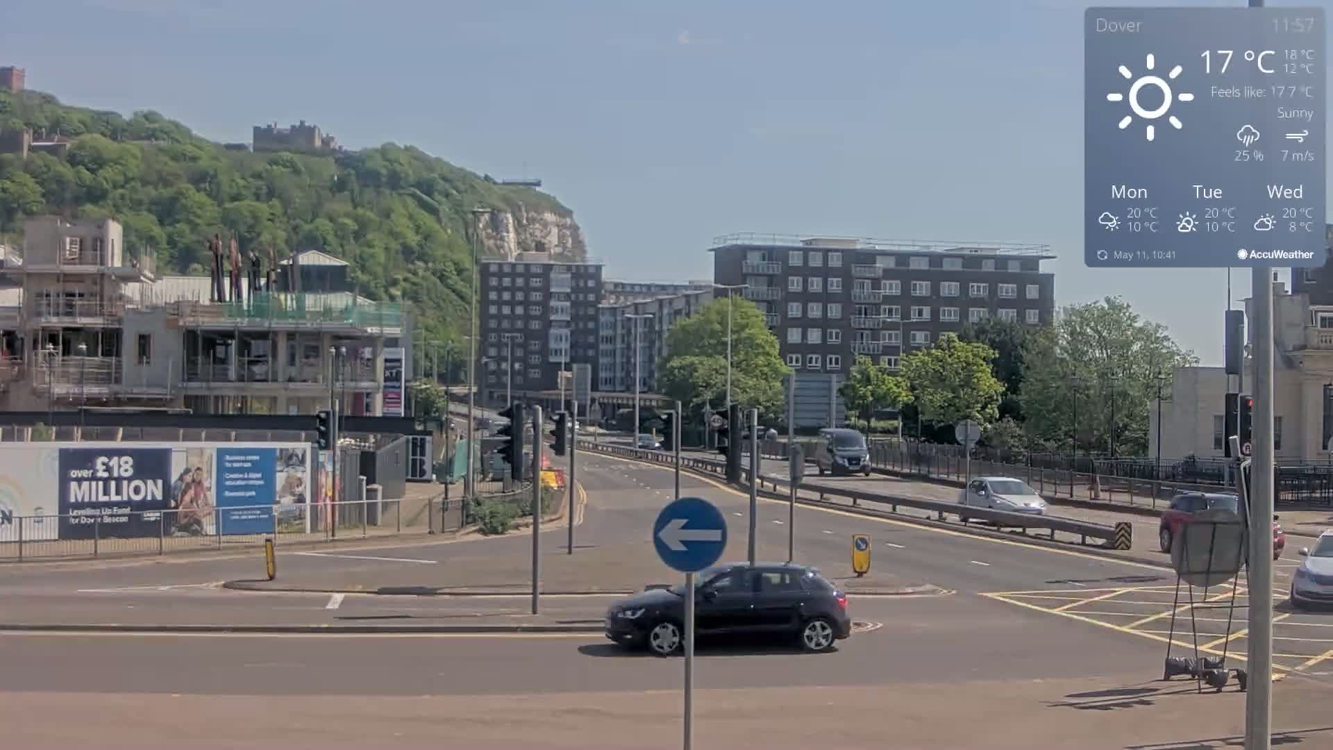 A sunny day reveals a city intersection with a black car turning left, several buildings, a construction site, and a hill with greenery and a castle in the background.