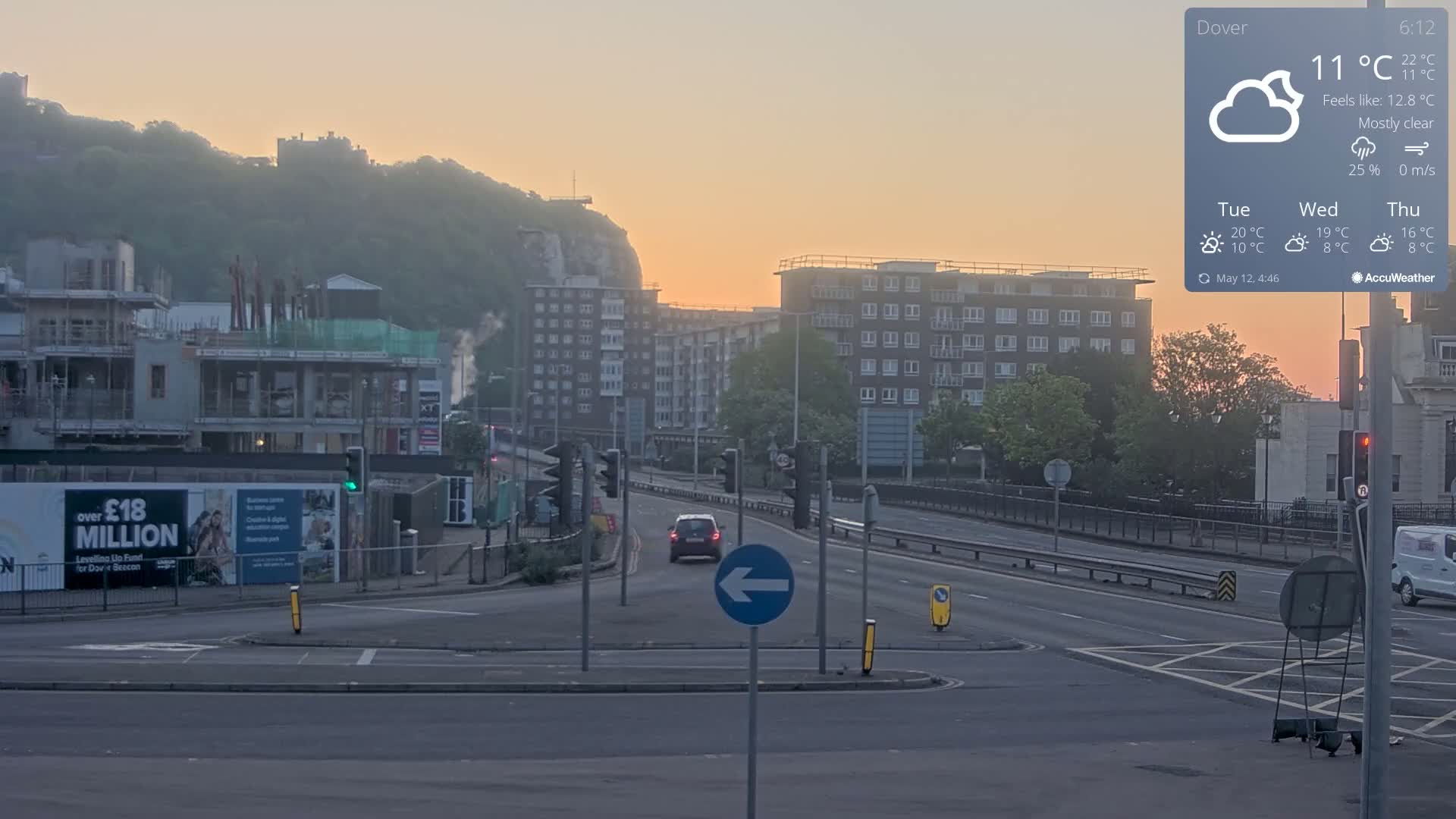 A mostly clear sunrise illuminates a city intersection with construction sites, apartment buildings, and a hill in the background.