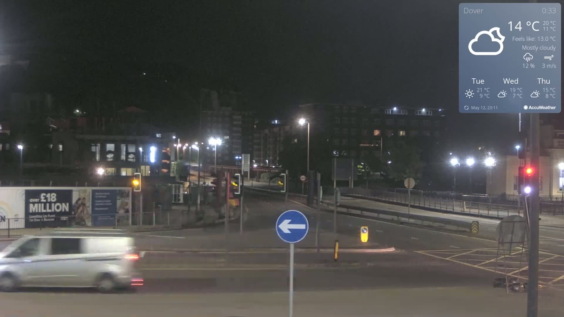A mostly cloudy night scene shows a street intersection with a white van driving by, streetlights illuminating buildings and a roadway.