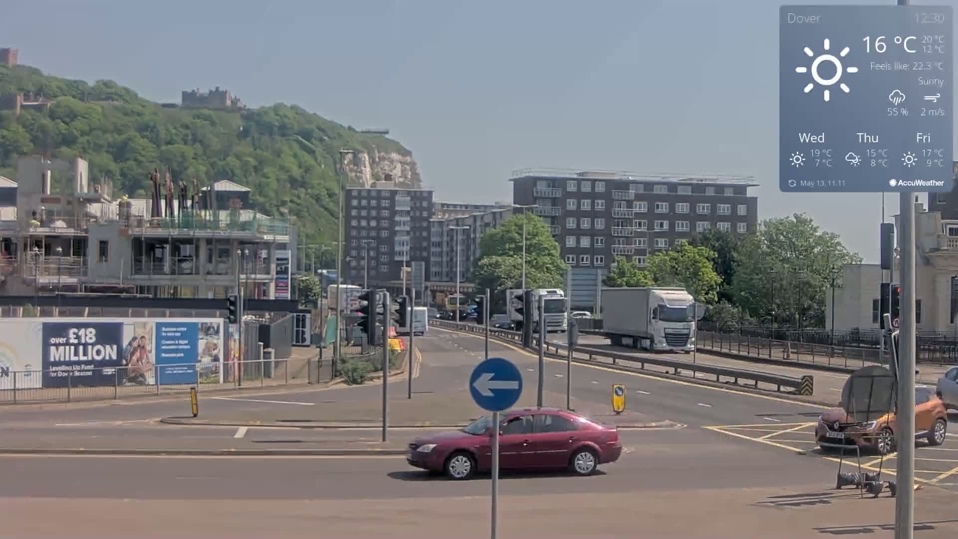 A sunny day reveals a multi-lane road intersection with several cars and trucks, buildings, and a distant hill with a castle under a clear sky.