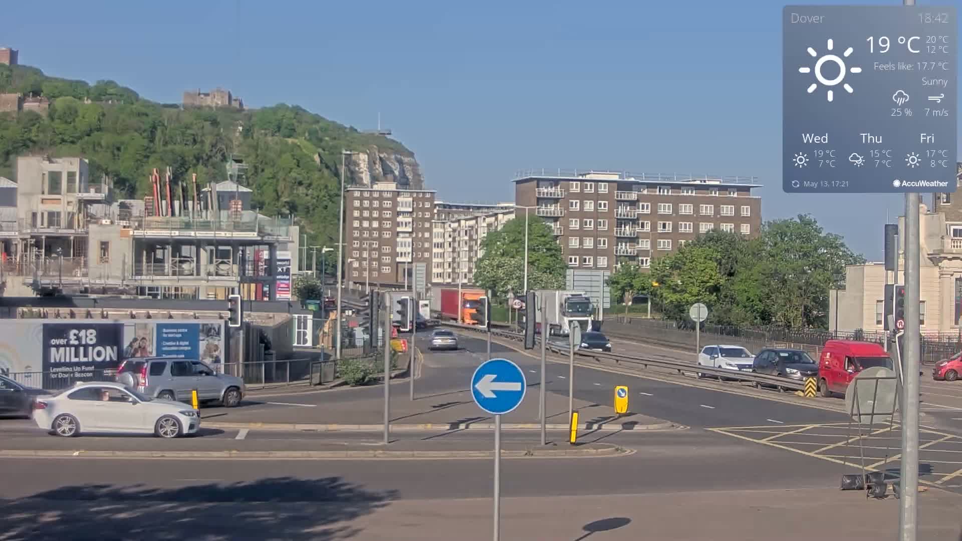 A sunny road intersection with several cars and trucks, a construction site, apartment buildings, and a hill with a castle in the background.