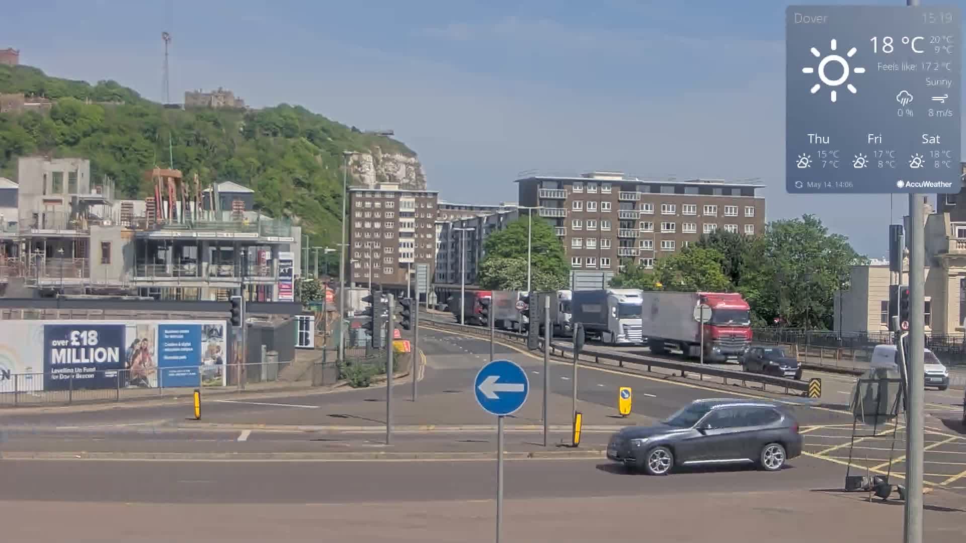 A sunny day reveals a busy road intersection near a construction site and apartment buildings, with a hill and castle visible in the background.