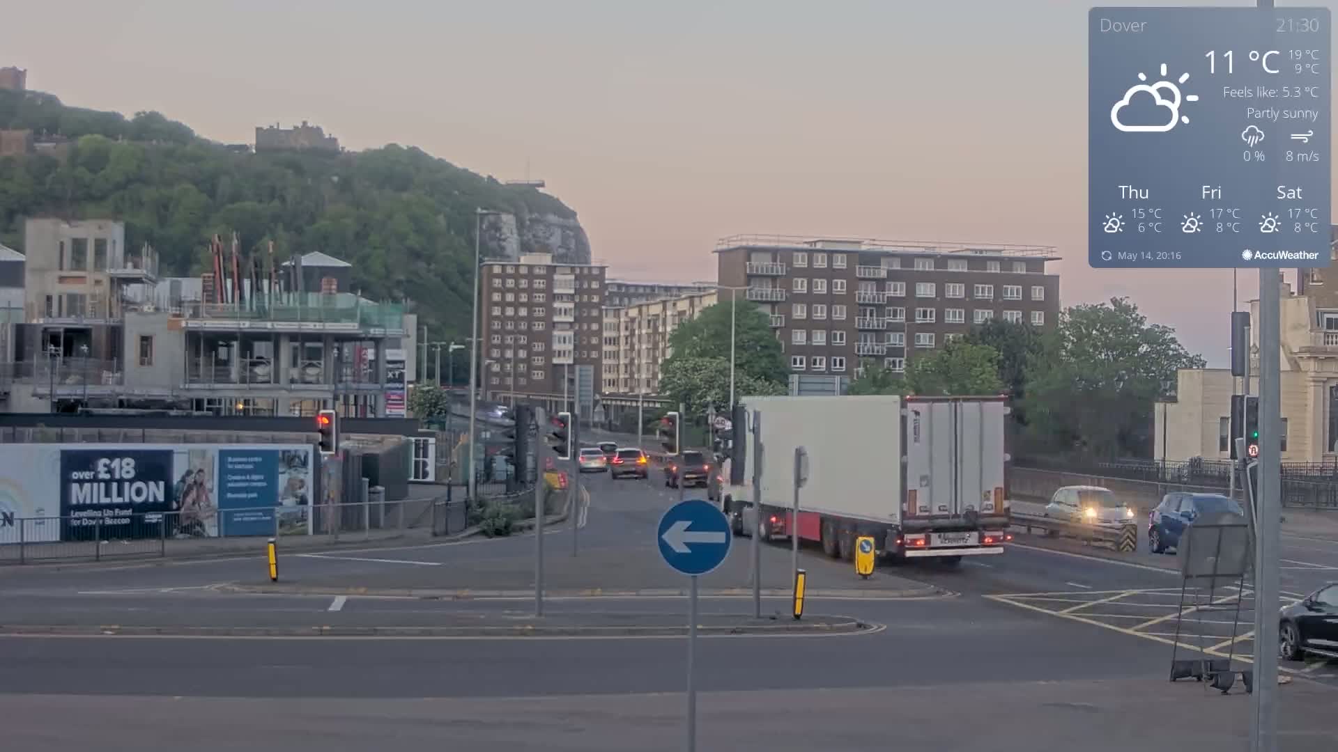 A partly sunny evening scene shows a multi-lane road intersection with several cars and a large white semi-truck, overlooked by apartment buildings and a hill with a castle on top.
