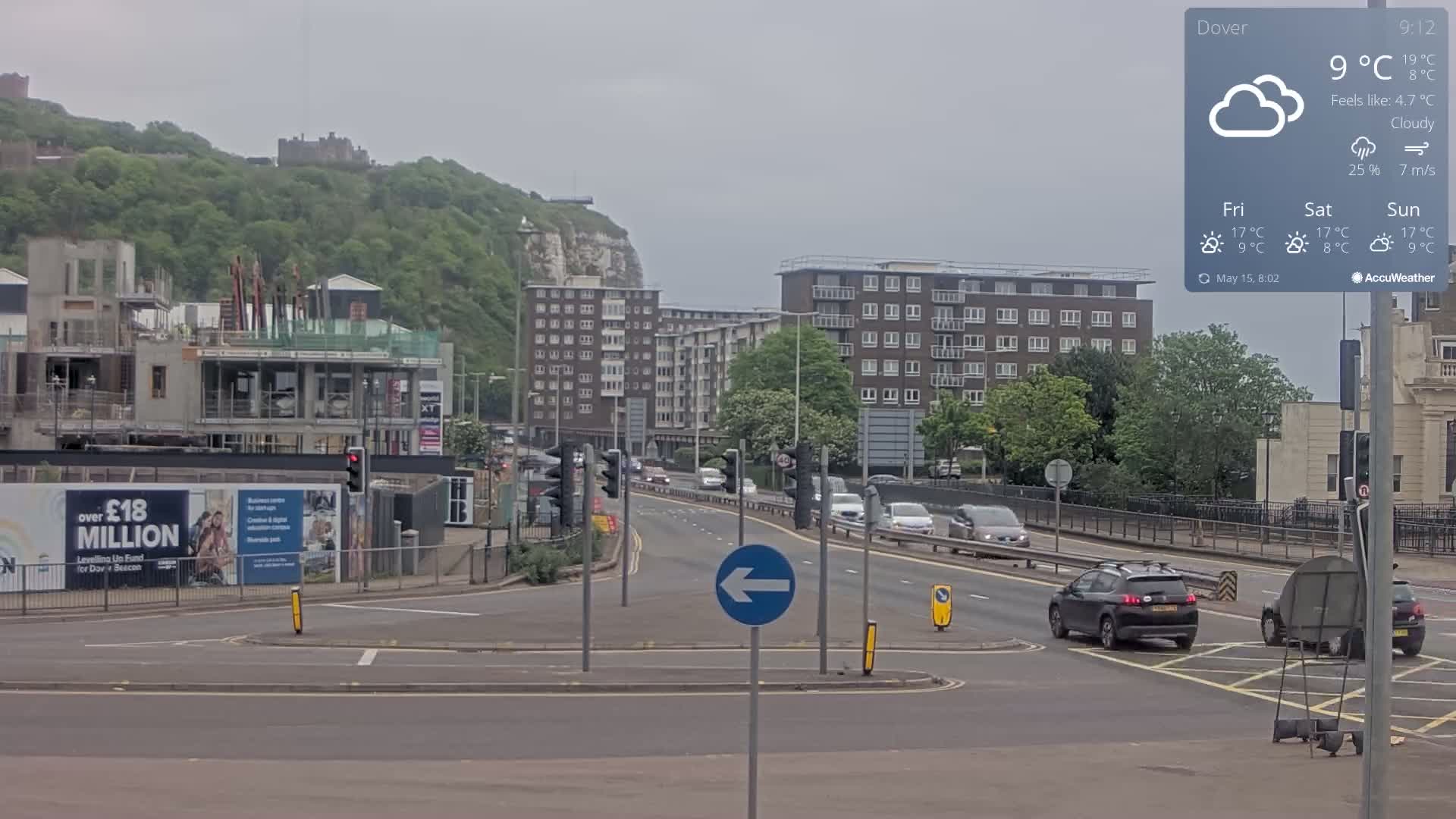 A cloudy day shows a multi-lane road with cars and buildings, including a partially constructed building, and a hill with a castle in the background.