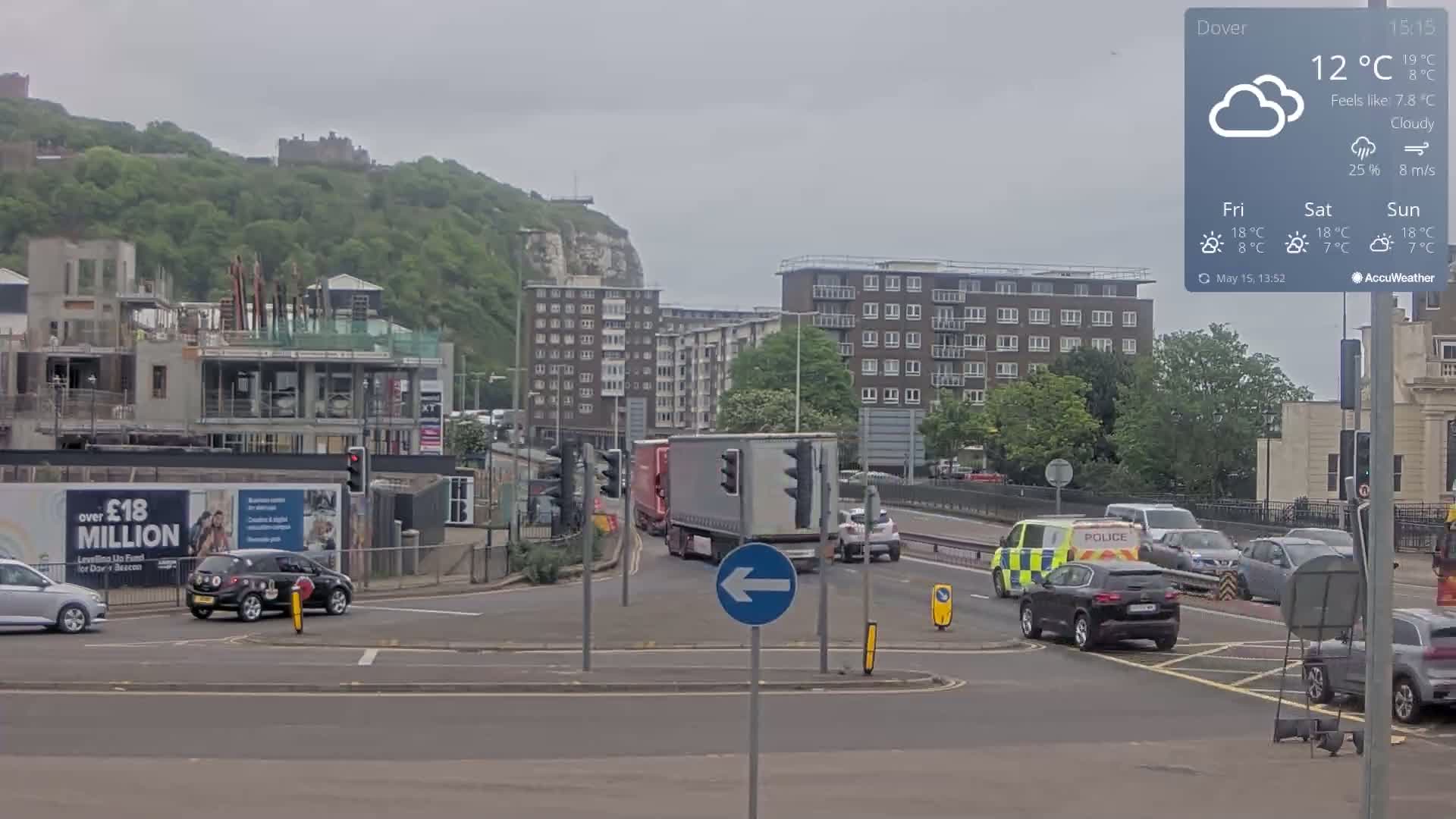 A cloudy, overcast day shows a traffic intersection with several vehicles, including a police van, near an apartment building and a partially constructed building, with a hill and castle visible in the background.
