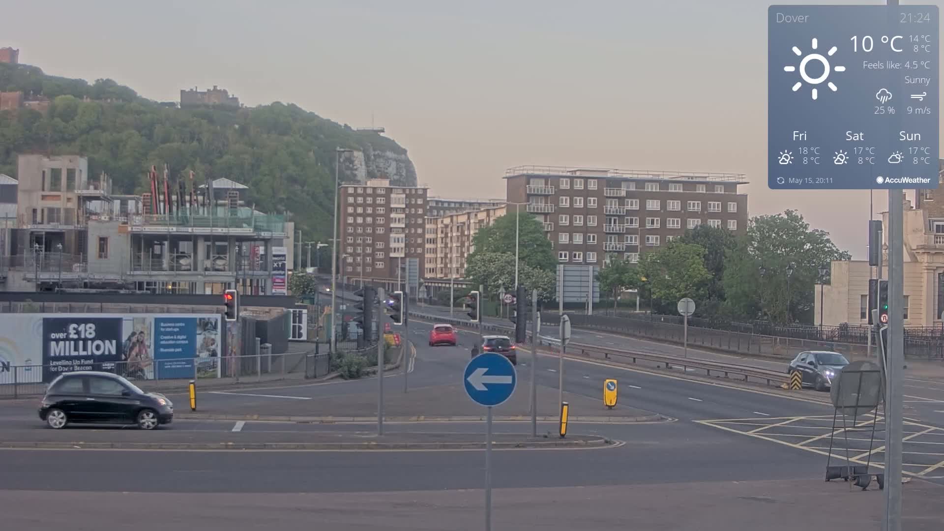 A sunny, but cool evening scene shows a multi-lane road with several cars, construction on the left, and apartment buildings and a castle atop a hill in the background.