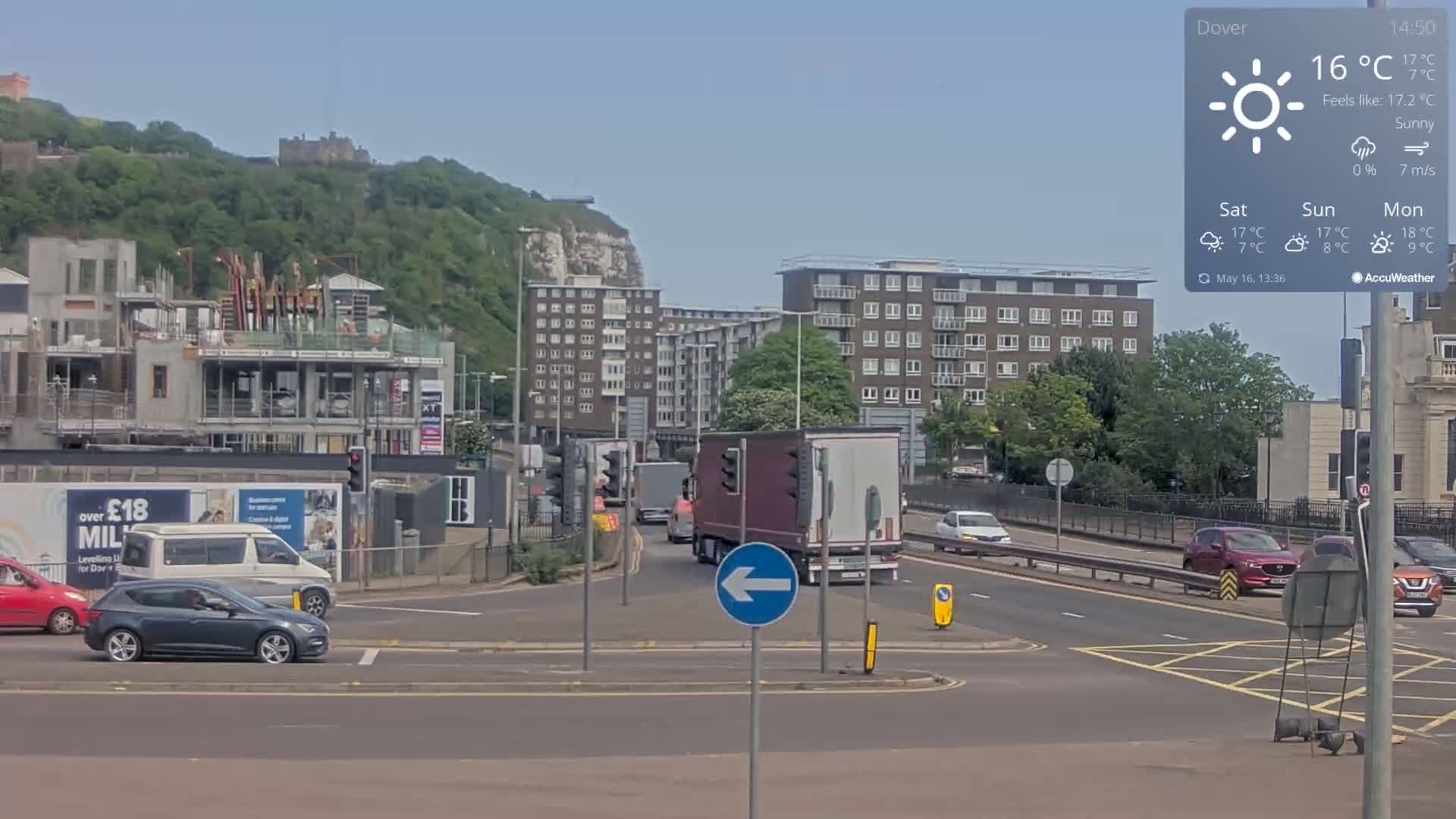 A sunny, daytime scene shows a multi-lane road intersection with several cars and trucks, a partially constructed building, residential buildings, and a hill in the background featuring a castle.