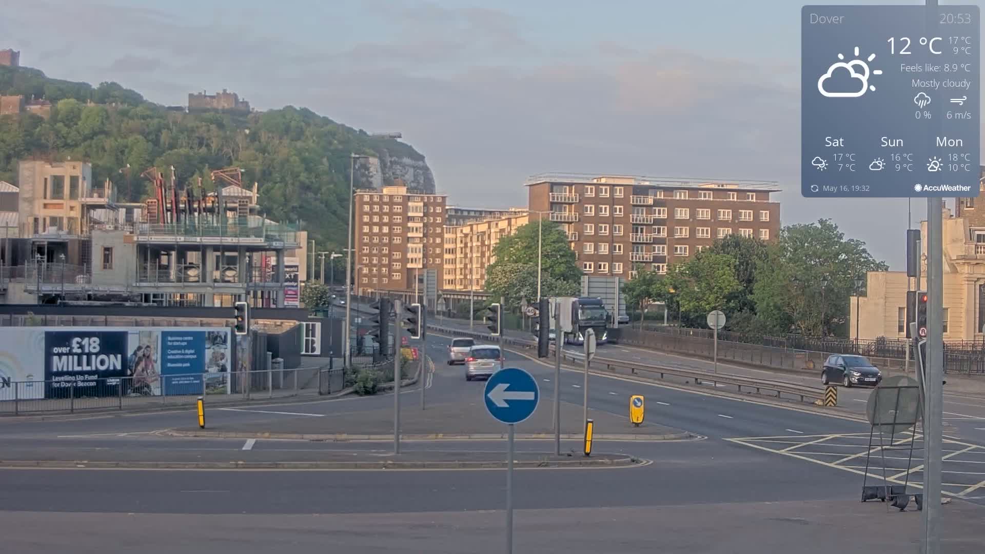 A mostly cloudy scene shows a roadway with several cars, a construction site, and apartment buildings, all set against a backdrop of a hill with a castle and white cliffs.