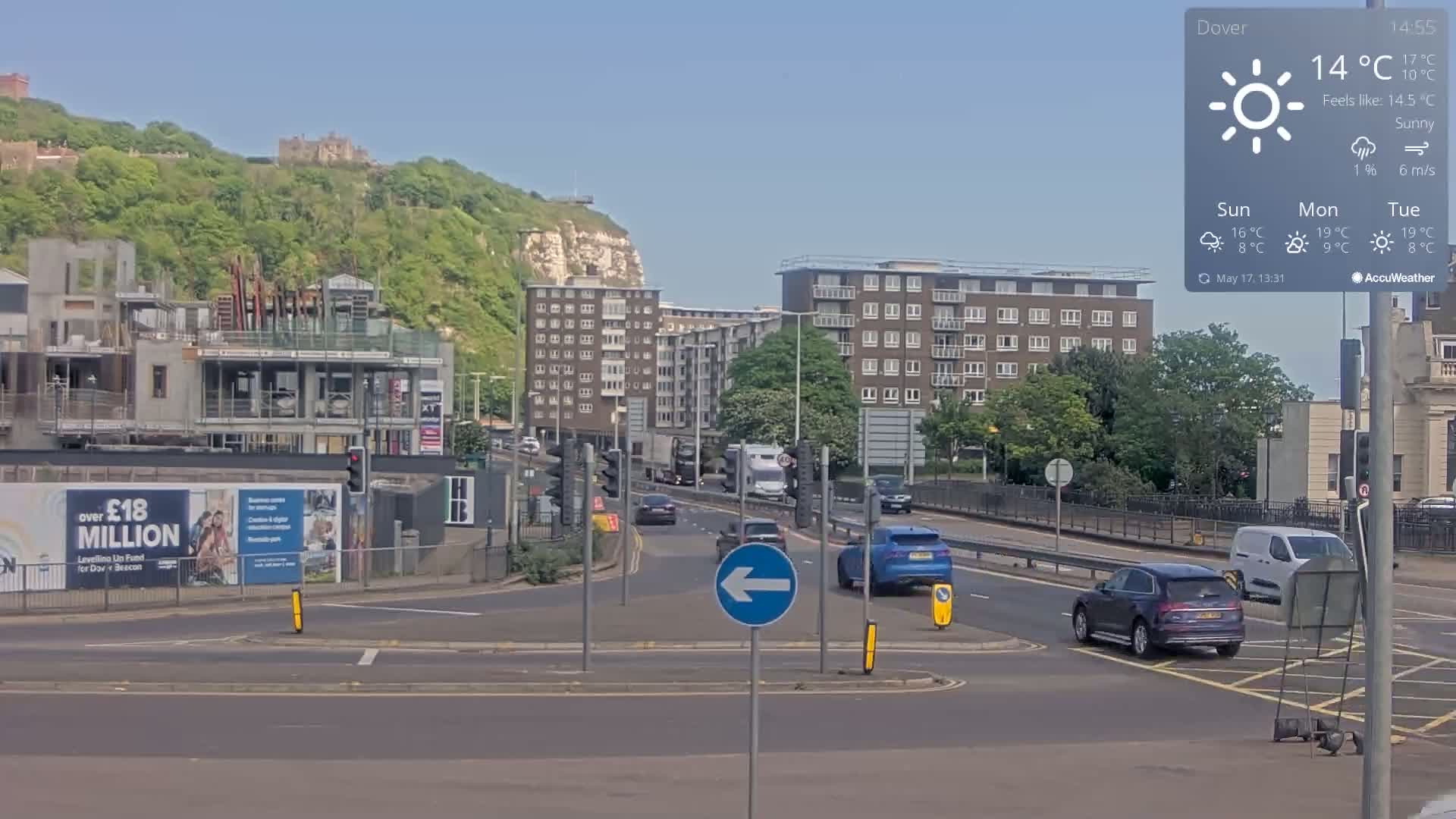 A sunny day shows a multi-lane road intersection with several cars, buildings, and a hill with lush greenery and a castle in the background.