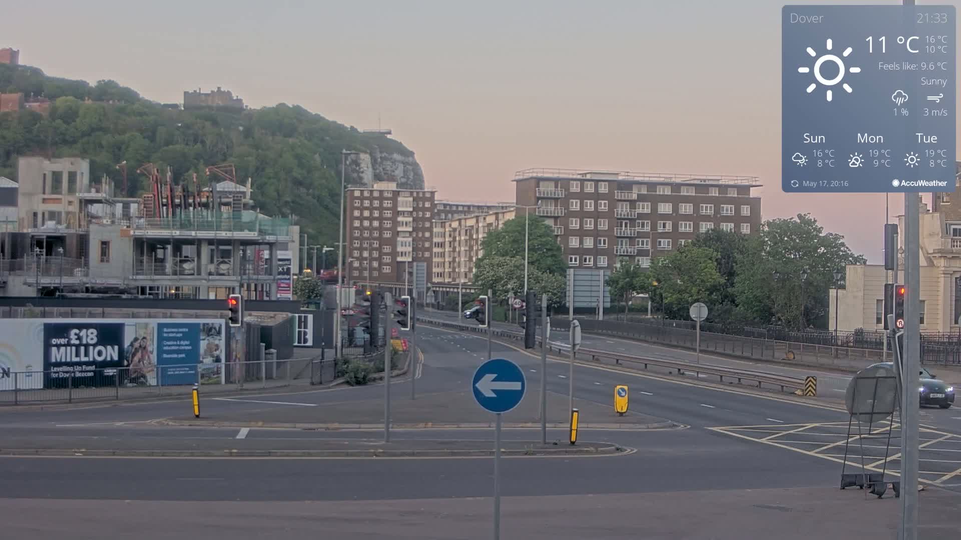 A mostly empty multi-lane road intersects with a roundabout, under a sunny sky, with apartment buildings and a construction site visible in the background, alongside a hill with greenery and a castle.
