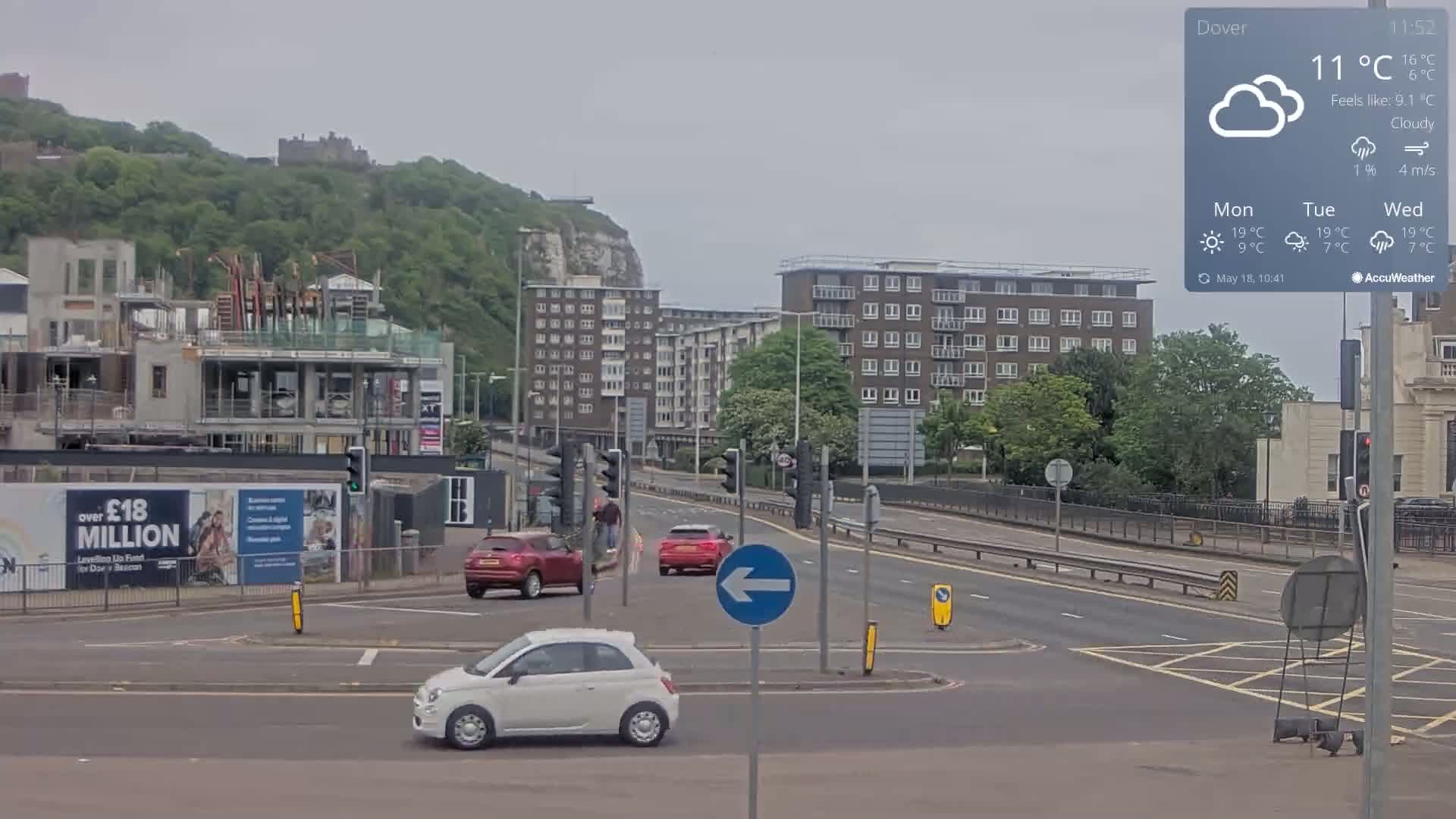 A cloudy day shows a busy intersection with several cars, buildings, and a partially constructed building near a hill with a castle in the background.